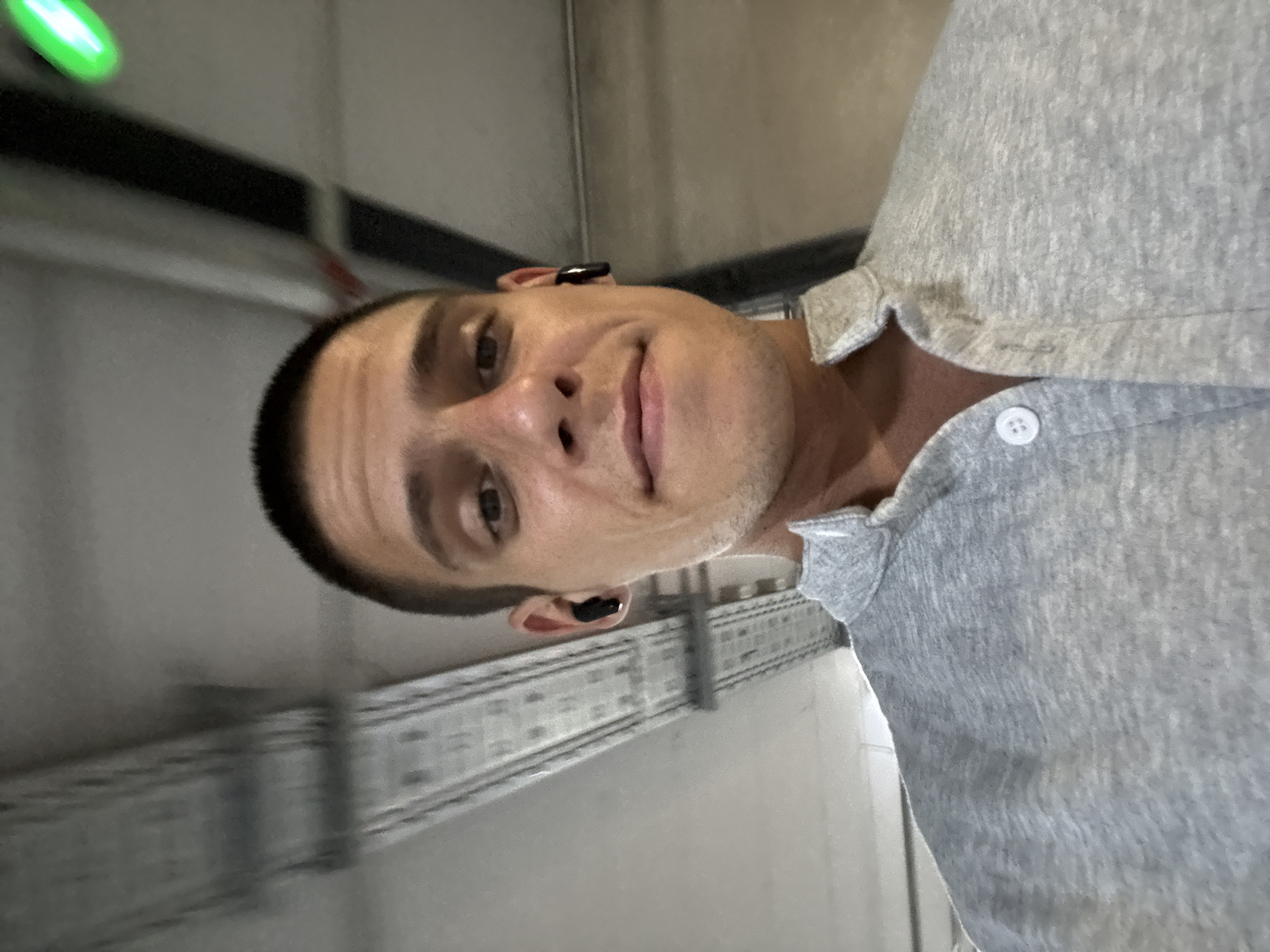 A young man with a buzzed haircut and a grey collared shirt is taking a selfie. He is wearing black wireless earbuds. The background appears to be an indoor parking garage with exposed pipes and lighting fixtures. A green light is visible in the upper right corner. The image is likely taken in Sofia, Bulgaria, based on the provided location.