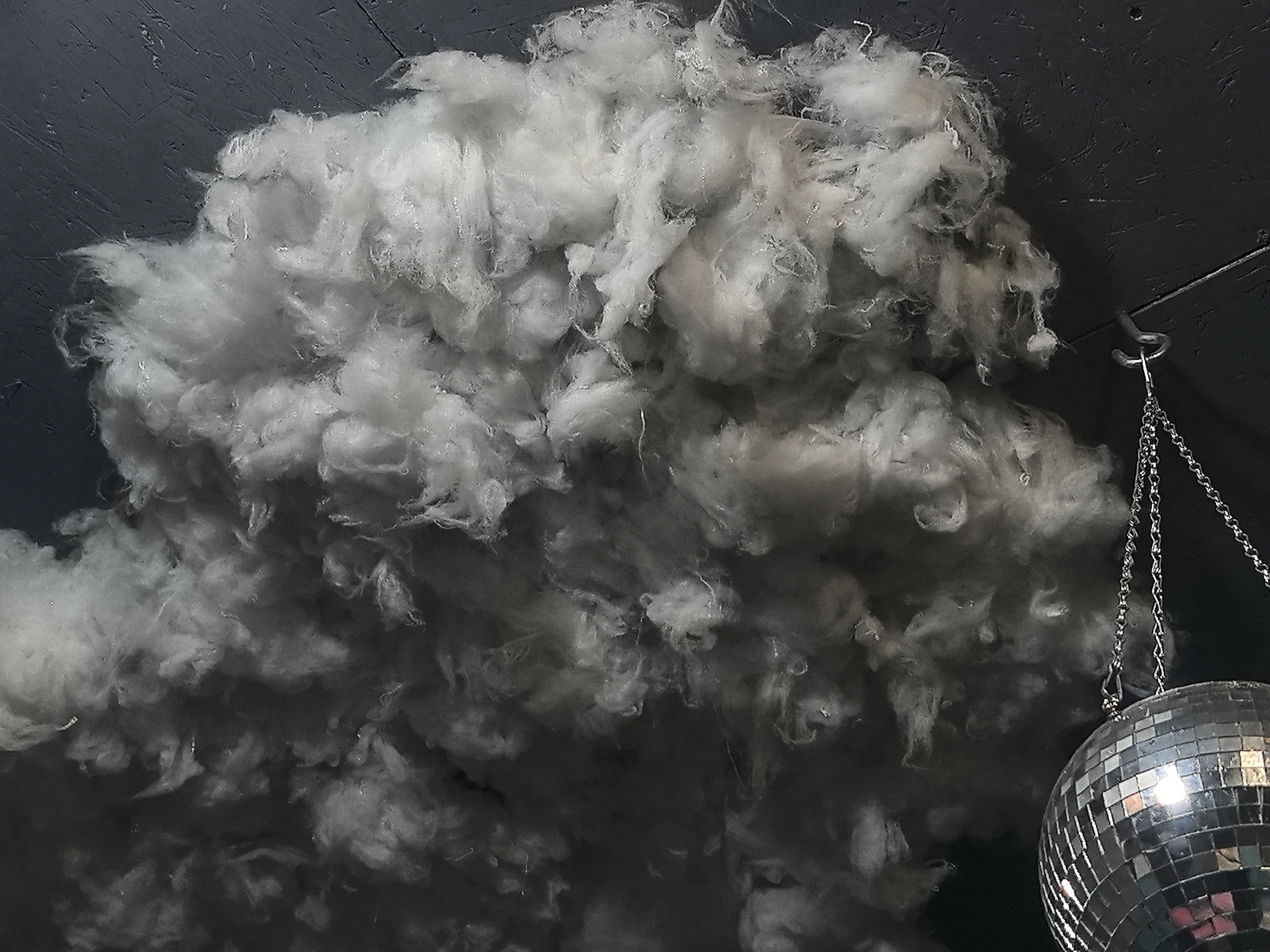 The image shows an interior with a large cloud-like mass of white, fluffy material attached to the ceiling. The ceiling is dark, possibly gray or black. A disco ball hangs from the ceiling, suspended by a chain. The overall lighting is dim, casting shadows and creating a dramatic effect. The location in Noviy, Russia, is not discernible from the image.