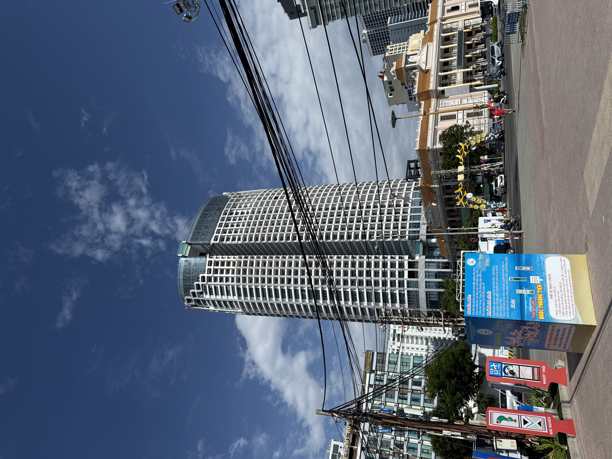 The image captures a street scene in Nha Trang, Vietnam, under a bright blue sky with scattered clouds. Dominating the background is a tall, modern skyscraper with a white facade featuring numerous rectangular windows, its curved design giving it a sleek, contemporary appearance. To the left of the skyscraper, other buildings of varying heights and architectural styles are visible, some appearing to be hotels or apartment complexes.
In the foreground, a large blue and yellow informational display stands on the sidewalk, featuring text in Vietnamese and images of products, possibly related to a promotion or event. Two red, triangular directional signs are placed to the left of the display, one pointing to "NHÌ NGHÊ" and the other to "NHƯỢT TRỜI." A tangle of black electrical wires crisscrosses the upper portion of the image, supported by wooden poles, a common sight in many urban areas.
The street itself is bustling with activity. Several motorcycles and cars are on the road, suggesting moderate traffic. A few individuals are visible walking or riding their motorcycles. In the mid-ground, a grand, older building with classical architecture, featuring columns and a light-colored facade with a reddish-brown roof, stands prominently. Decorative yellow archways, possibly for a festival or event, are erected in front of this building. The ground in the foreground is paved with reddish-brown square tiles. The overall impression is of a vibrant urban environment in Vietnam.