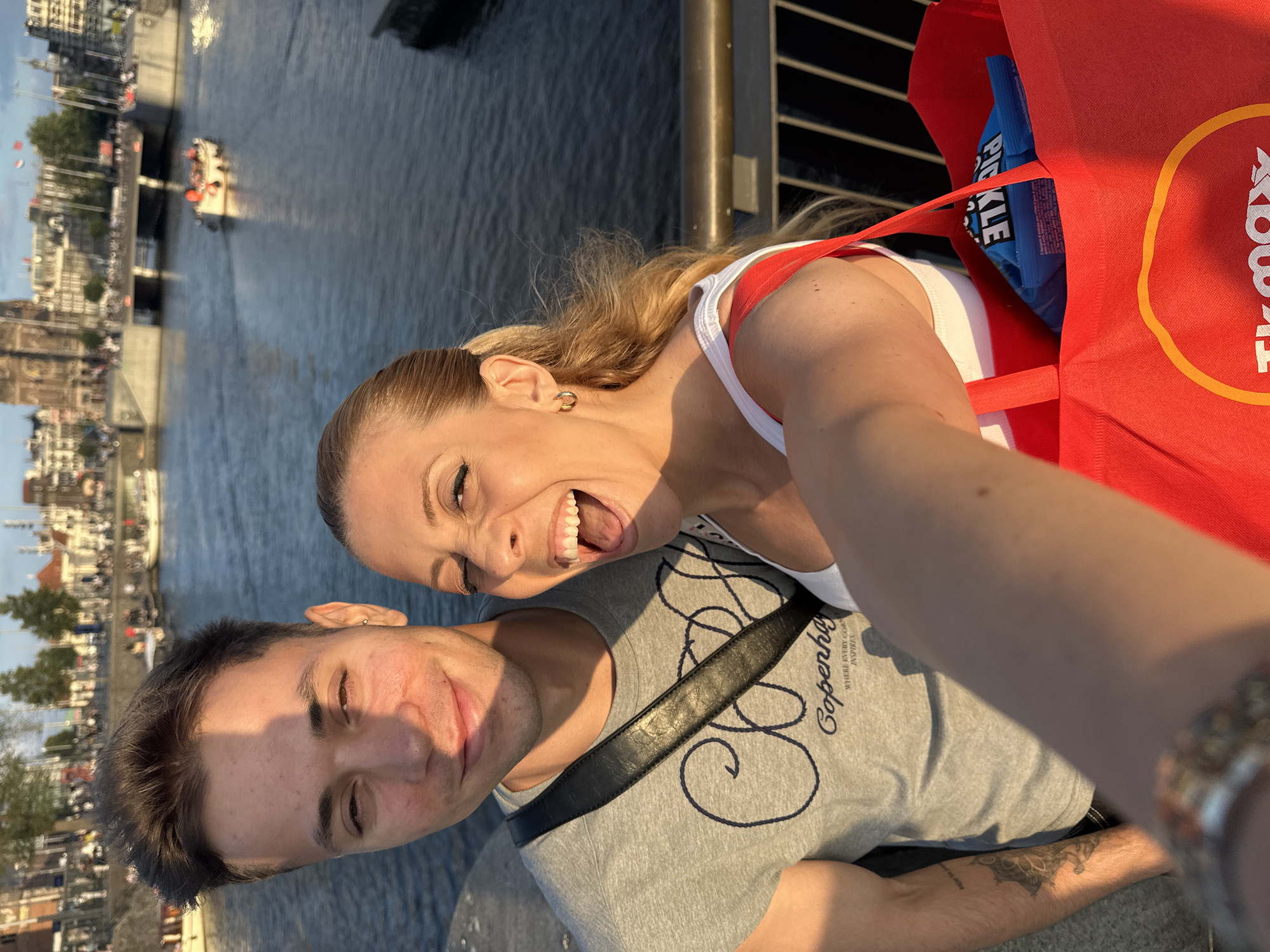 A selfie is taken by a couple on a bridge overlooking a canal in Amsterdam, Netherlands. The man in the foreground on the left is wearing a grey t-shirt with the word "Copenhagen" printed on it. He has short brown hair and is looking towards the camera with a slight smile. The woman on the right is wearing a white tank top and has blonde hair pulled back in a ponytail. She is laughing with her mouth open, looking at the camera. She is holding a red shopping bag with the TK Maxx logo visible. Behind them, a canal with boats and buildings typical of Amsterdam line the waterway. The sun is casting long shadows, indicating it is likely late afternoon or early evening.