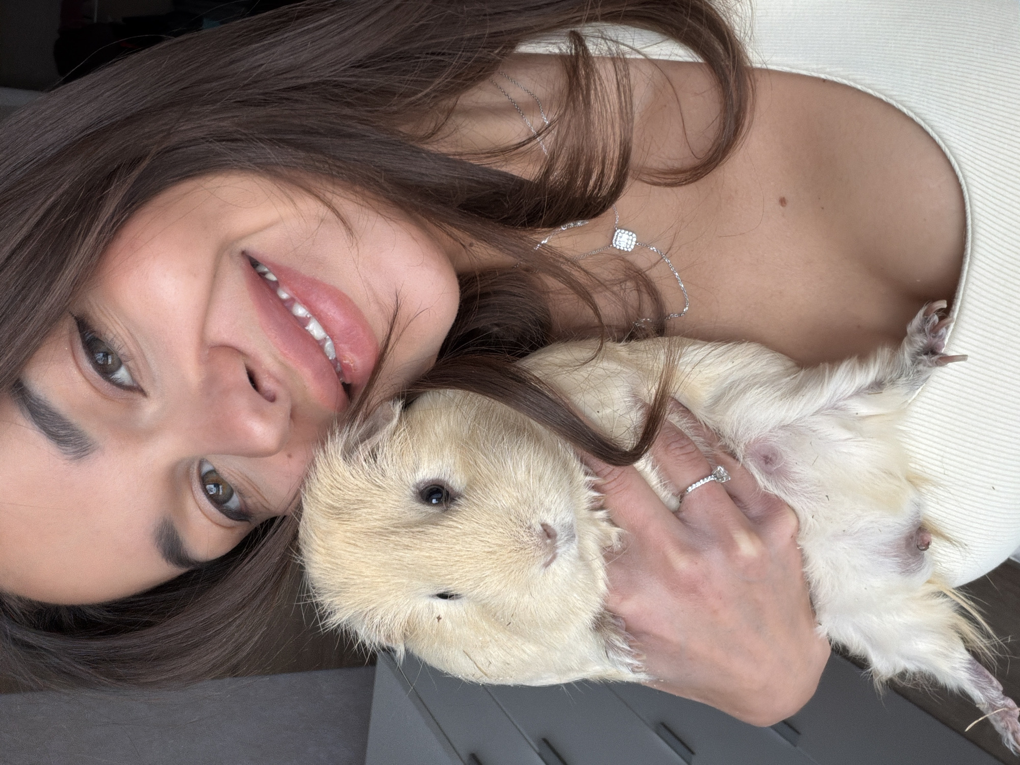 A woman is holding a light brown and white guinea pig. The woman is fair-skinned with brown hair. She is smiling and wearing a necklace. The guinea pig is being held close to her chest. A ring is visible on the woman's finger. The background is a grey surface. The location is not identifiable as Krasnoyarsk, Russia.