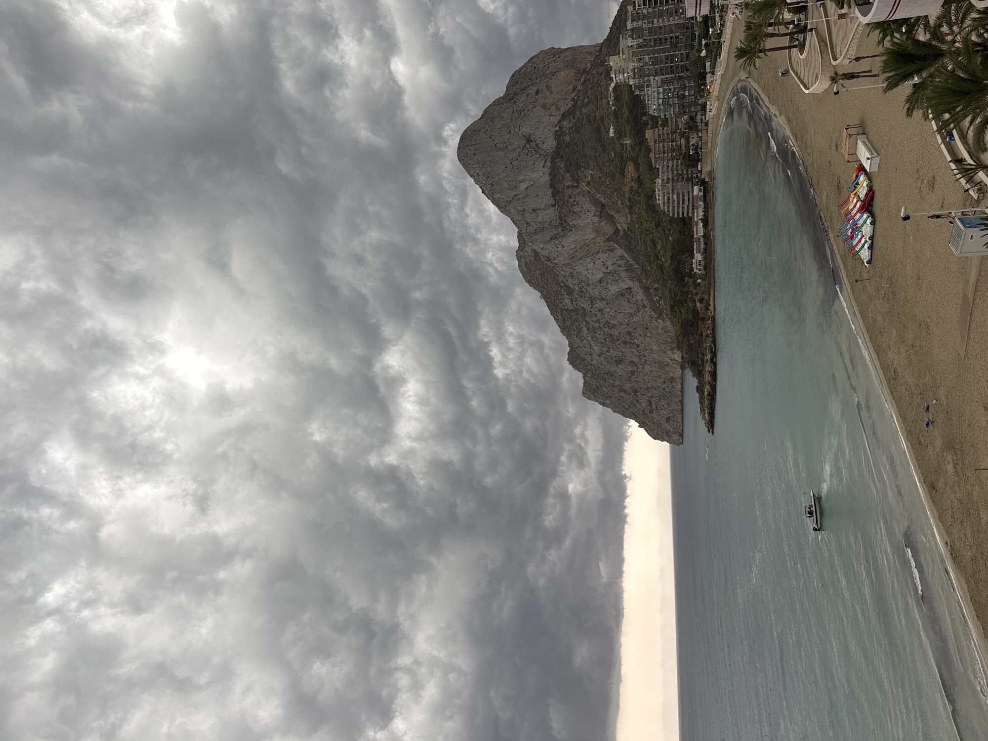 This is an exterior, eye-level, medium shot of the coastline of Calp, Spain, under a cloudy sky. The focus is on a large, rocky mountain that juts out into the sea, with a sandy beach and several buildings in the foreground. The sea is calm and a light turquoise color, with small waves lapping at the shore. A white boat with a red stripe is visible in the water, with several people on board. The beach is mostly empty, with a few scattered beach chairs and a cluster of colorful pedal boats near the water's edge. The buildings in the background are modern high-rises, suggesting a developed tourist area. The overall impression is one of a slightly overcast day at the beach, with the imposing presence of the mountain dominating the landscape.