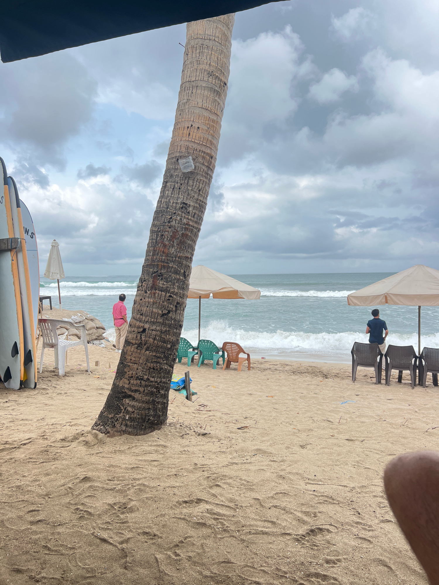 A medium shot captures a beach scene in Kuta, Indonesia, under an overcast sky. In the foreground, the textured bark of a large palm tree dominates the left side of the frame. The sand is light brown and appears undisturbed in many areas, with some footprints visible. To the left of the palm tree, several surfboards are propped up, leaning against a wooden structure.
Beyond the surfboards, a white plastic chair sits on the sand. Further back, a man in a pink shirt and beige pants stands facing the ocean. He is positioned between a stack of sandbags and a white umbrella. The ocean stretches to the horizon, with moderate waves breaking on the shore, creating white foam.
To the right of the frame, two beige beach umbrellas provide shade over several plastic chairs. One umbrella is positioned further back than the other. Beneath the front umbrella, a man in a dark blue shirt and light-colored shorts stands facing the ocean. In front of him are several dark plastic chairs. To the left of these chairs, a few green plastic chairs are visible, and to the right, more dark plastic chairs are set up. A blue floatation device is visible on the sand near the base of a wooden pole. The overall impression is a slightly cloudy day at a popular beach destination.