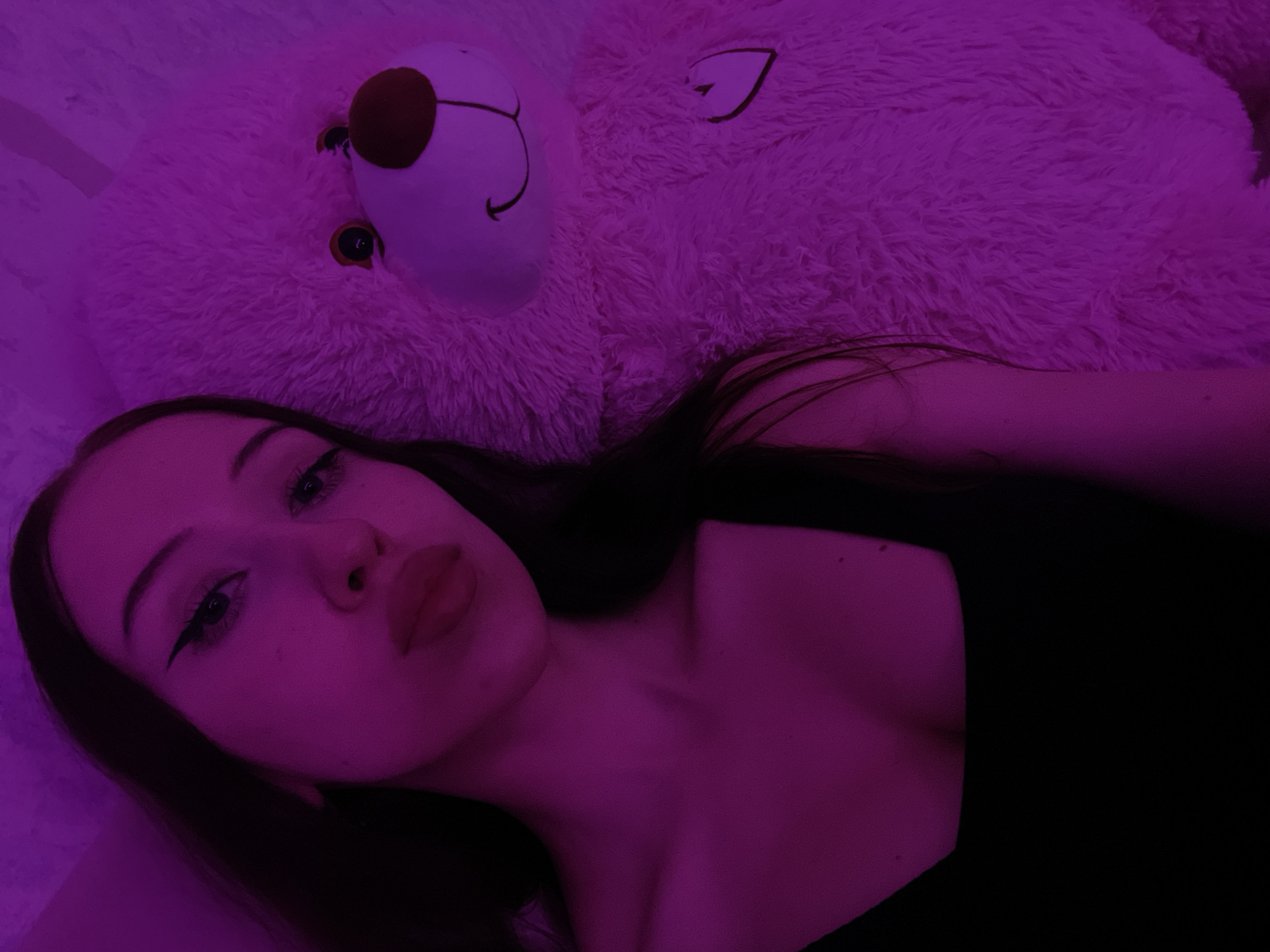 The image is a close-up, waist-up shot of a young woman with long dark hair and fair skin, taken indoors under purple lighting. She is wearing a black, square-necked top and is positioned to the left of the frame, leaning against a large, fluffy pink teddy bear. The teddy bear is on the right side of the frame and appears to be soft and plush. The woman's face is partially visible, with her eyes looking towards the camera and her lips slightly pursed. Her makeup includes winged eyeliner and lipstick. The background is a textured wall, also illuminated by the purple light, giving the scene a somewhat intimate and atmospheric feel. There is no discernible indication of the city of Votkinsk, Russia, in the image itself.