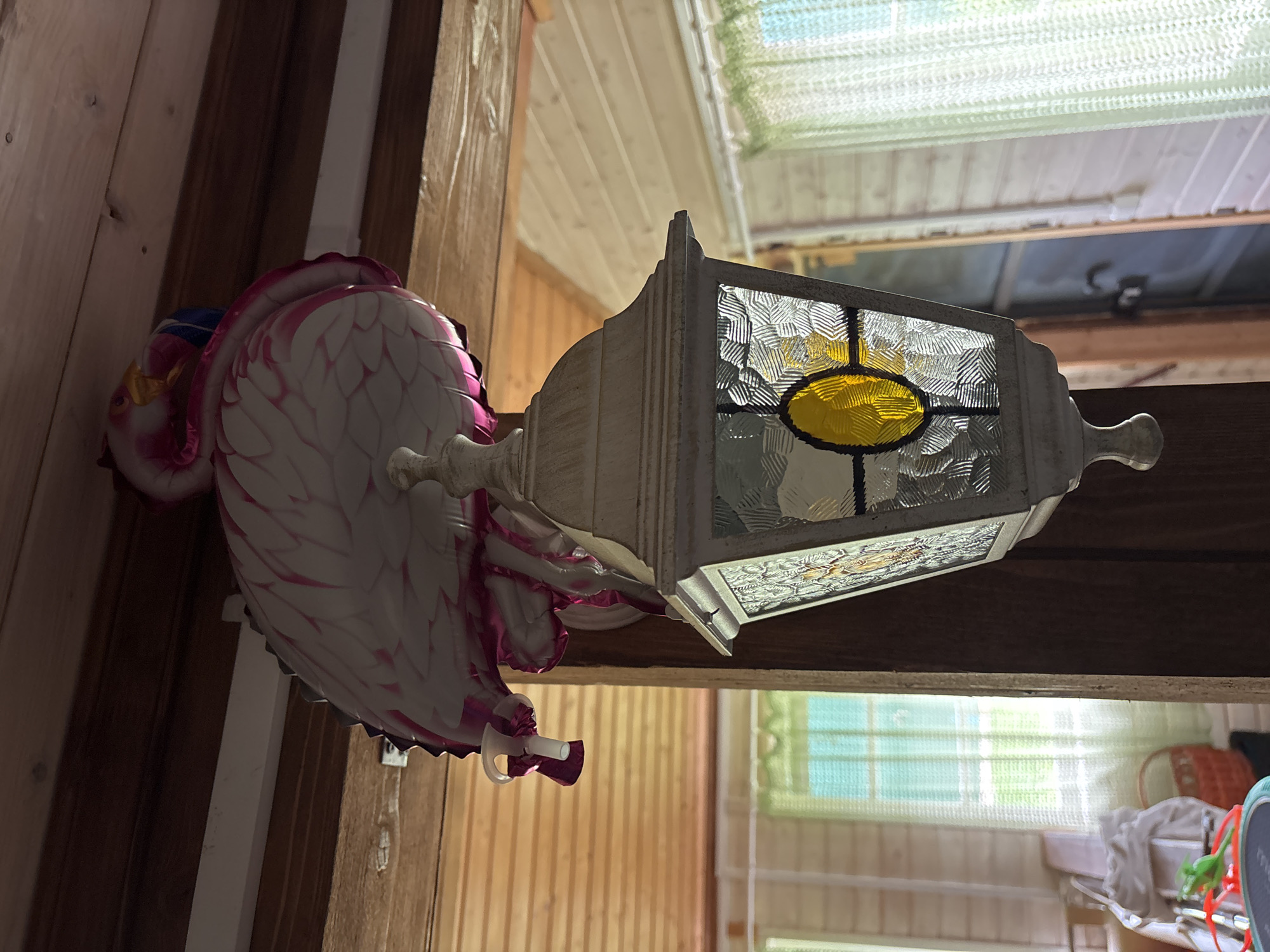 A decorative flamingo ornament is perched above a white outdoor lantern with stained glass panels. The lantern is attached to a wooden beam, which is part of the ceiling of what appears to be a rustic wooden interior. The walls of the interior are also made of wood planks, and two windows with light green curtains are visible in the background. The overall scene suggests an indoor setting in a wooden structure, possibly a house or a dacha, in the city of Borki, Russia. The lighting is somewhat dim, with the natural light from the windows illuminating the space. No people are present in the image.