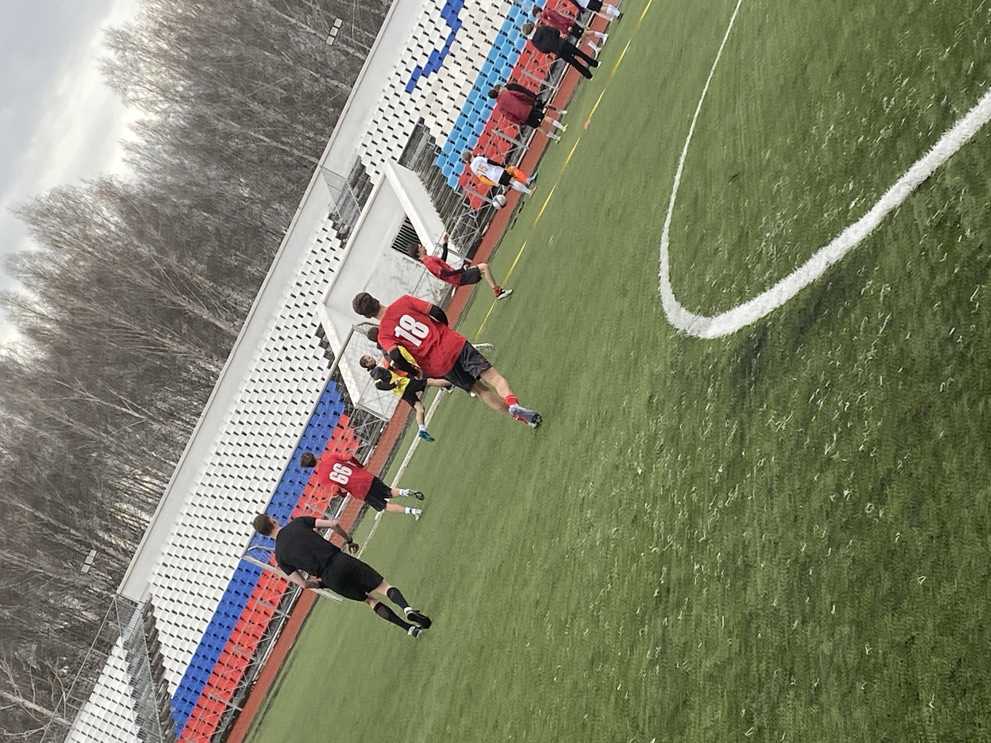 The image shows a soccer field in Chaikovsky, Russia. A soccer team is practicing on the field, wearing red jerseys with numbers on the back. The field has white lines marking boundaries and a grassy surface. A grandstand with red, white, and blue seats is located near the field. Bare trees stand in the background, and the sky is overcast.
