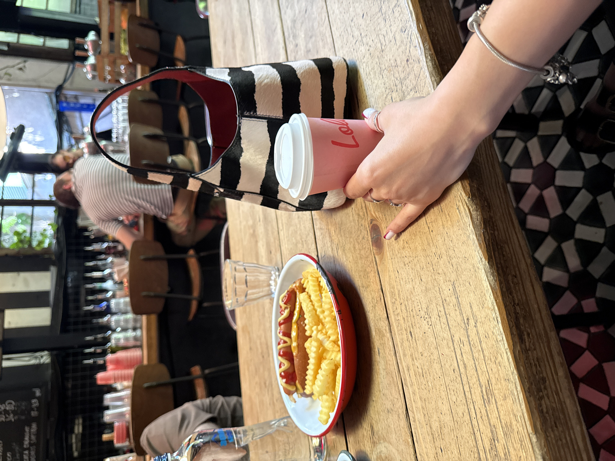 The image depicts a close-up view of a wooden table in what appears to be a casual establishment in Tbilisi, Georgia. In the foreground, a person's hand, adorned with rings and a bracelet with charms, is holding a pink disposable coffee cup with red lettering. To the left, a red and white enamel plate contains a hot dog topped with ketchup and mustard, served alongside a generous portion of crinkle-cut french fries. A clear glass with ribbed detailing is situated behind the plate. In the background, a black and white striped handbag rests on the table, and further back, a bar area is visible with bottles on shelves and people seated at stools. The overall atmosphere suggests a relaxed, urban dining experience.