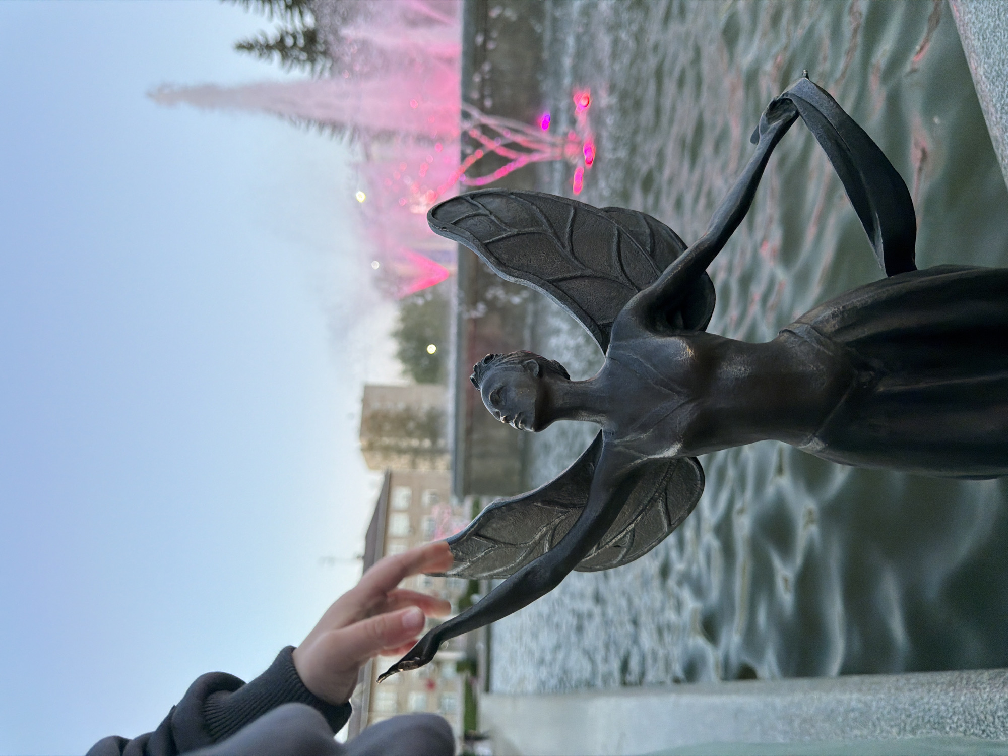 A bronze statue of an angel with wings is the focal point, positioned in front of a fountain with illuminated pink water jets. A hand is reaching towards the statue. The statue is positioned on a grey stone surface. Behind the fountain, there are buildings. The location is Chaikovsky, Russia.