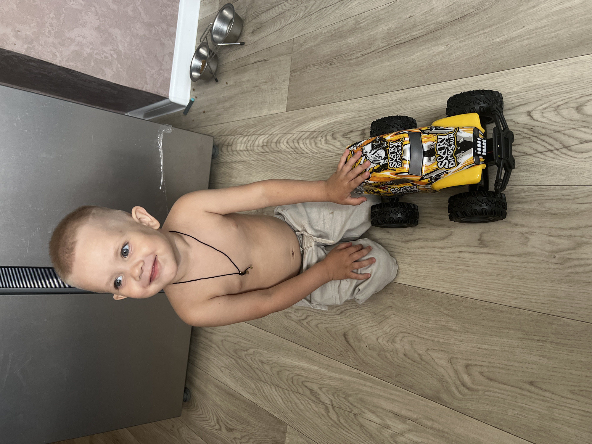 A young boy, likely a toddler, is sitting on a light-colored wooden floor next to a metallic refrigerator. He is shirtless, wearing light-colored pants, and has a small black necklace with a charm. He is looking up and smiling directly at the camera, with a happy and playful expression. His right hand rests on his knee, while his left hand is placed on a yellow and black toy car.
The toy car is an RC car with large, rugged tires. It has a graphic design featuring skulls and the words "SCARY DINOSAUR" printed on its body. The car is positioned to the right of the boy.
In the background, above the refrigerator, are two metal pet bowls on a stand, suggesting the presence of pets. The lighting indicates it is daytime, and the scene appears to be indoors in a domestic setting, possibly a kitchen or dining area, in Марковский, Russia. The overall mood is cheerful and candid.