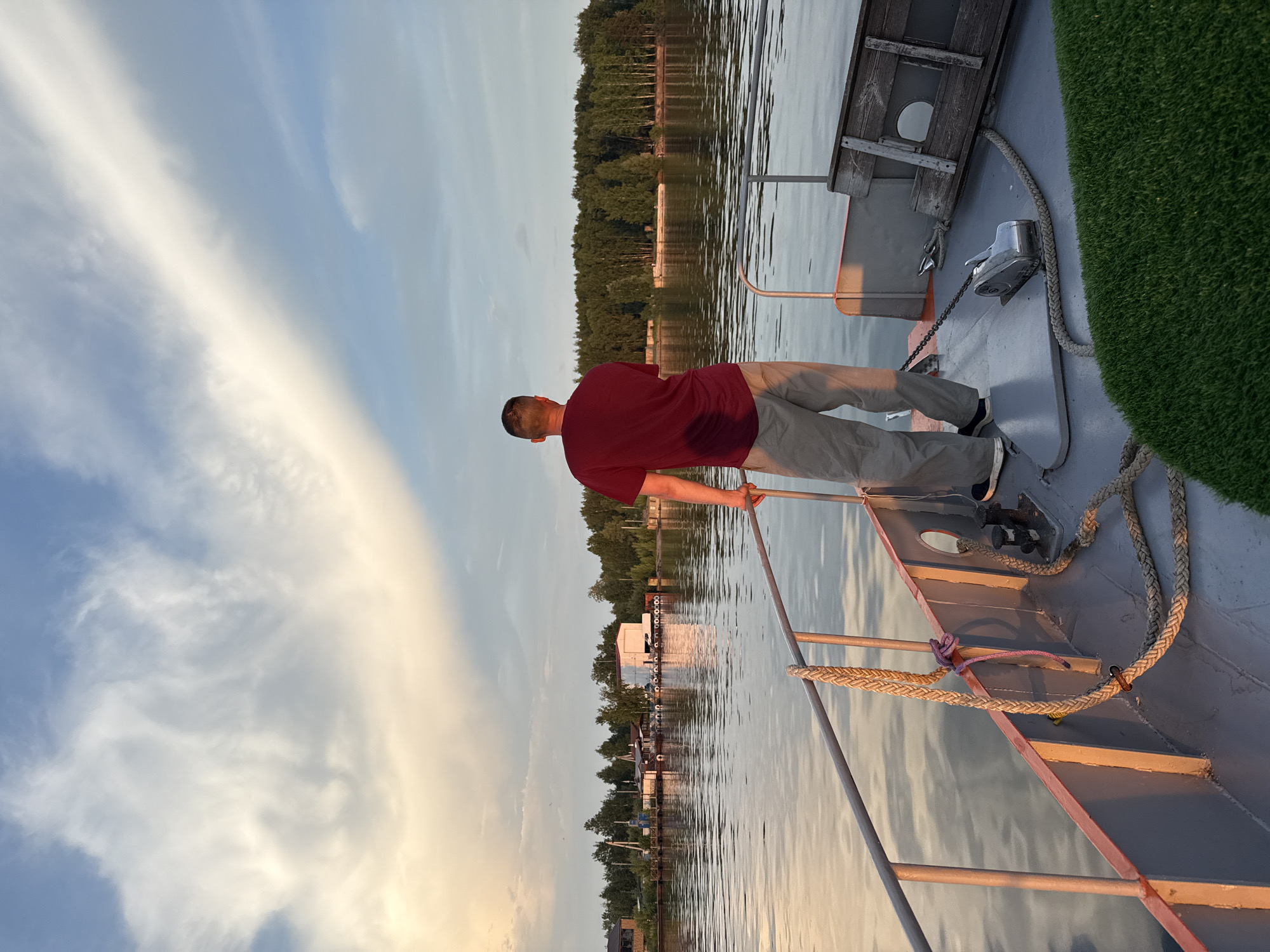 A man stands on the deck of a boat, facing away from the camera and looking out at a body of water. He is wearing a dark red t-shirt and light gray pants. The boat has a railing and several ropes secured to it. In the background, across the water, there is a shoreline with trees and several buildings, including what appear to be houses and possibly some industrial structures. The sky above is a mix of blue and white clouds, with a soft, warm light suggesting either sunrise or sunset. The water is calm, reflecting the sky and the shoreline. The scene is likely taking place on the Kama River near the city of Chaikovsky, Russia, given the context provided.