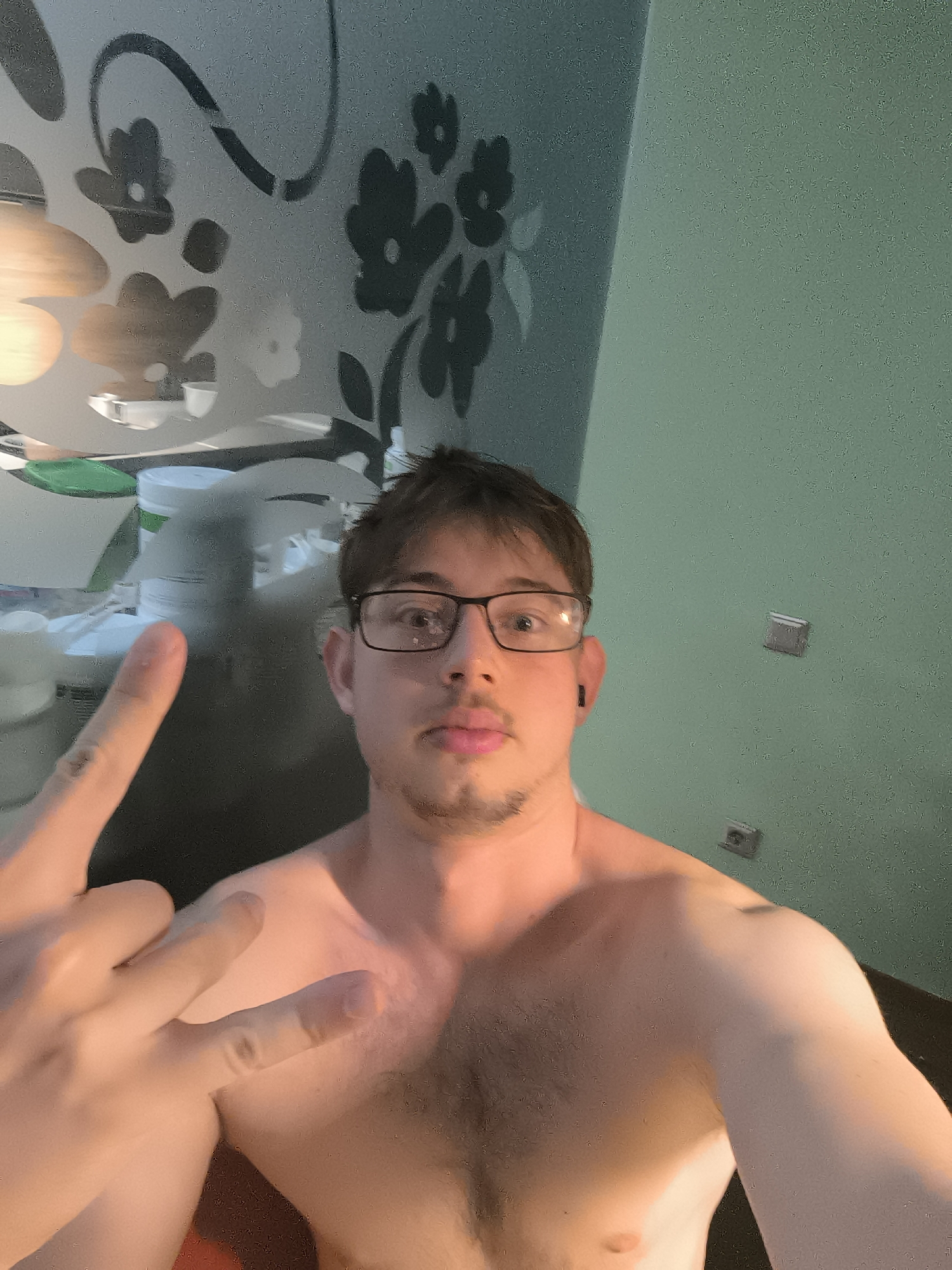 A young adult male, bare-chested and with dark hair, takes a close-up selfie indoors, possibly in Sofia, Bulgaria. He wears black-rimmed glasses and a small black earbud in his right ear, with a short beard or goatee. His left hand is raised towards the camera, forming a gesture with the index and middle fingers extended upwards, and the thumb and pinky also extended, resembling a "rock on" or "peace" sign. His expression is neutral with a slight pout.
The background on the left side features a frosted glass partition or door decorated with a dark, stylized floral pattern of flowers and swirling lines. Behind this glass, several out-of-focus objects, including a large white container (possibly a supplement jar), are visible, suggesting a counter or shelf. The right side of the background shows a plain, muted teal-green wall, on which two light grey electrical outlets or switches are mounted. The lighting is artificial, casting a warm glow on his right shoulder and chest. The setting appears to be a casual home interior, possibly a bathroom or a room adjacent to one. No visible text is present.