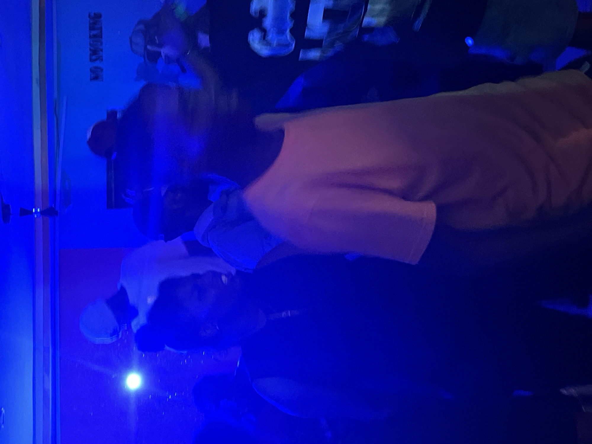 In Durumi, Nigeria, a group of individuals is gathered in a dimly lit room, illuminated by vibrant blue lighting. The scene appears to be a casual social event, possibly a party or gathering. In the foreground, a person in a black sleeveless shirt is facing left, with a faint smile. To their right, another individual in an orange shirt is seen from the side, facing away from the camera. In the background, the wall displays a "NO SMOKING" sign, hinting at a regulated space. Other individuals are present, but their features are obscured by the low light and their movement. The overall atmosphere is energetic and festive, with a focus on the social interaction of the people present.