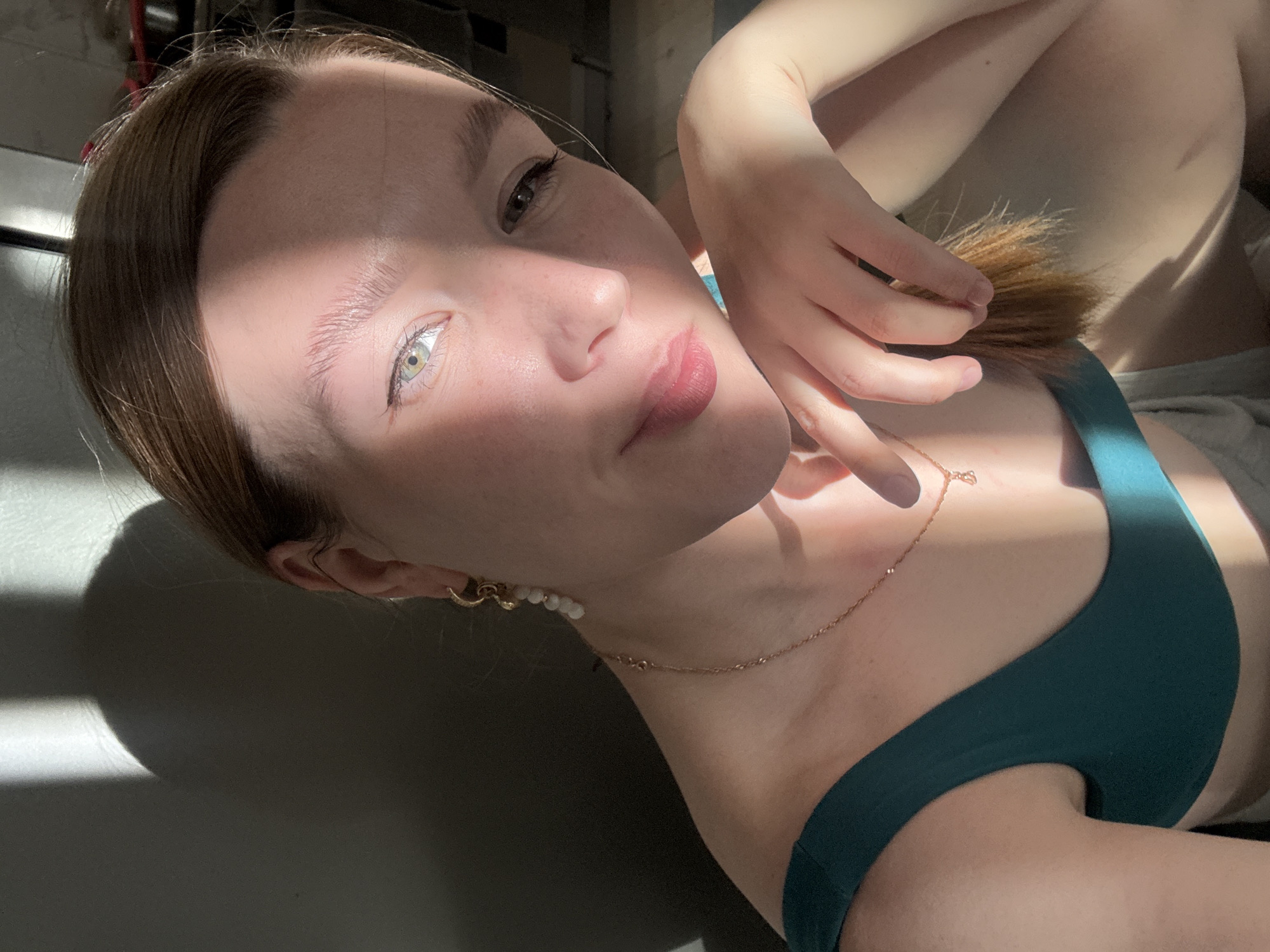 The image is an angled, close-up selfie of a young woman lying down indoors in Izhevsk, Russia. The main subject is a woman with light brown hair, fair skin, and light green or hazel eyes. She is looking directly into the camera with a soft, slightly smiling expression. Her lips are a natural pink tone.
The setting appears to be a brightly lit room, with strong, natural sunlight casting distinct, angled shadows across her face and the surrounding surfaces, suggesting either early morning or late afternoon. The light highlights parts of her face and body while leaving other areas in shadow.
She is wearing a dark teal or green top, possibly a bralette or camisole. Accessories include a delicate gold chain necklace with a small pendant and a gold hoop earring on her visible left ear, adorned with small white, pearl-like beads. Her right hand is gently positioned near her cheek and neck, with her fingers slightly curled. The background is mostly dark and indistinct, offering no specific details about the room other than it being an indoor environment with significant natural light. There is no visible text in the image.