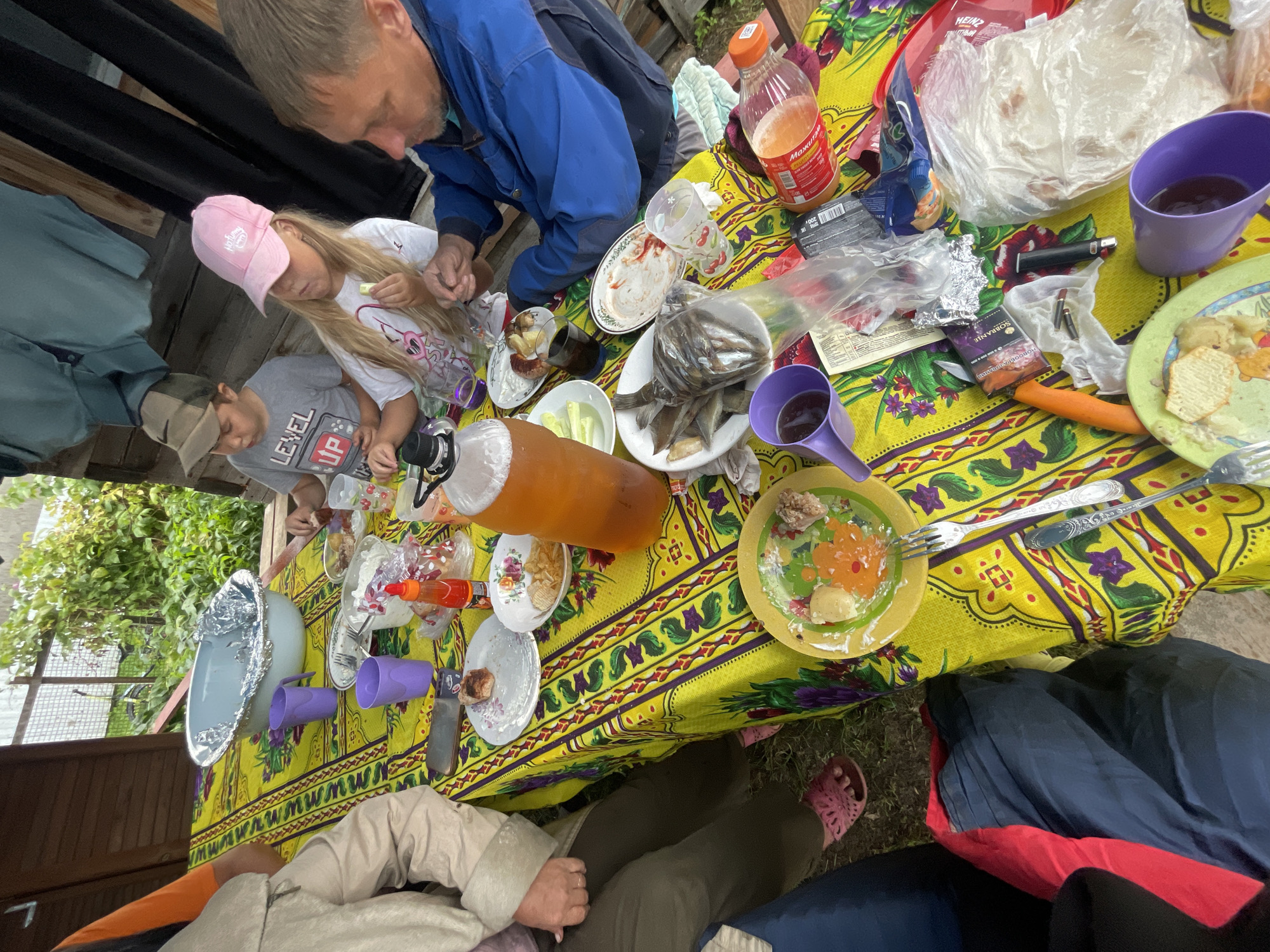 In Kamenka, Russia, a group of individuals, including at least two children and one adult male, are gathered around a brightly colored, patterned tablecloth laid out on a table, likely outdoors under a covered structure. The table is laden with food and drinks, suggesting a meal or picnic. Dishes visible include what appears to be fried fish, potato chips, a piece of meat on a plate, and various other food items in bowls and on plates. Beverages include a large bottle of amber-colored liquid, a bottle of red drink, and several cups, some purple. A child wearing a pink cap and a boy in a grey t-shirt with a cap are actively eating. The adult male, wearing a blue jacket, is also present at the table. The background shows some greenery and a wooden structure, consistent with an outdoor setting in a semi-rural or suburban environment. Various disposable items like plastic cutlery, plastic bags, and a lighter are also scattered on the table. The overall scene depicts a casual gathering and meal.