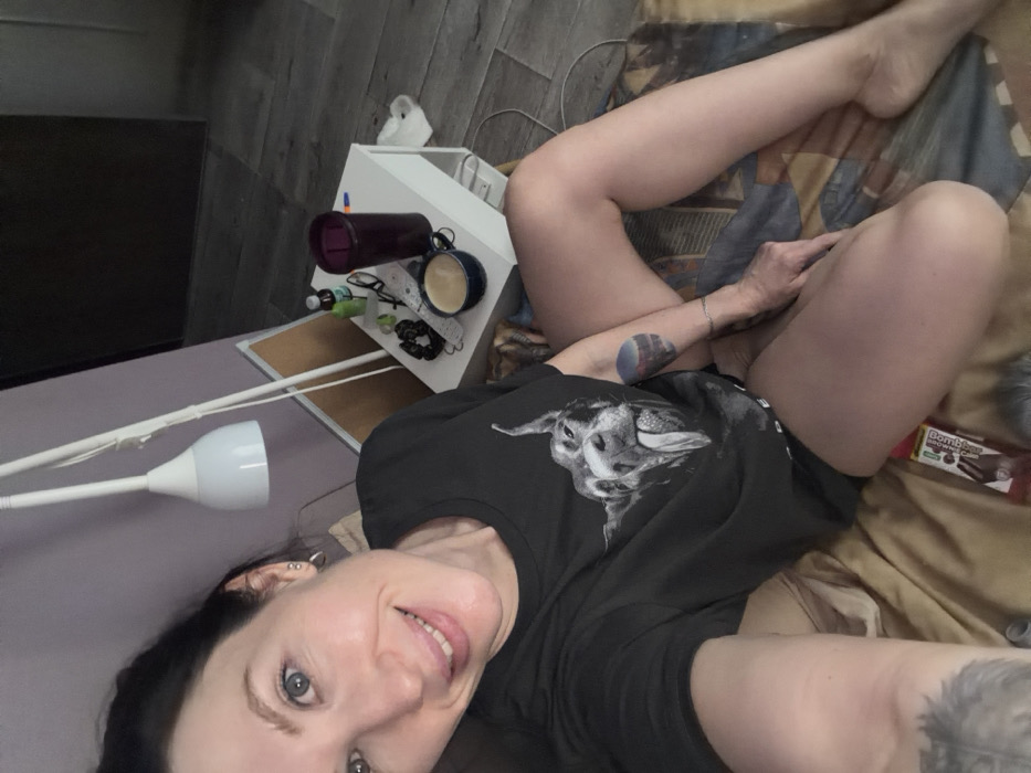 A female individual, appearing to be in her 30s or 40s with dark hair and blue eyes, is depicted in an indoor residential setting in Perm, Russia, taking a selfie. She is smiling directly at the camera, showing a slight toothy grin, and wears a black short-sleeved t-shirt featuring a greyscale graphic of a dog with its tongue extended. Visible tattoos include a circular, multi-colored landscape design on her left forearm and a darker, less distinct tattoo on her right forearm. She is seated or reclining on patterned bedding, with bare legs partially visible. To her right, a white bedside table holds a dark purple lidded tumbler, a blue ceramic mug containing a light-colored beverage with foam, black-framed glasses, several black hair ties, small bottles, and a white remote control. A white modern floor lamp with a conical shade extends over the scene, positioned against a light purple wall. Grey-toned wood-look flooring is visible in the background. A red and white packaged snack bar is present on the bedding near her right thigh.