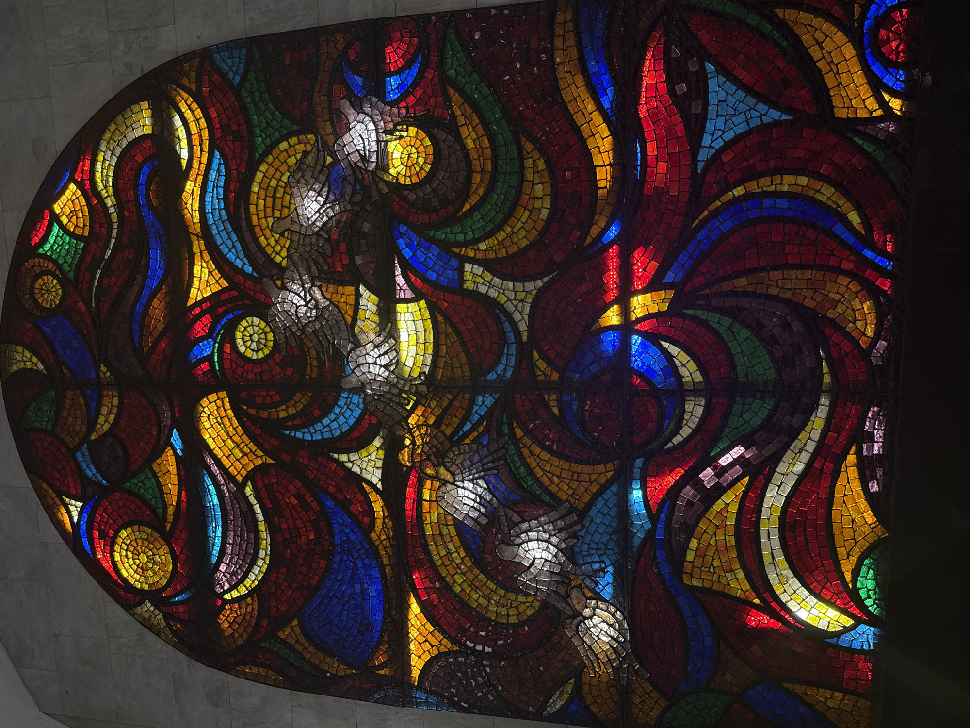 The image shows a large, colorful stained glass mosaic. The mosaic is predominantly red, yellow, and blue, with swirls of other colors like green, orange, and brown throughout. The overall design consists of curving, flame-like shapes, and circular patterns. In the central area, there are stylized white figures resembling birds, or perhaps angels. The mosaic is backlit, causing the colors to glow. The mosaic is set into a curved wall. To the left of the mosaic, is a light gray stone. This mosaic is located in the Novoslobodskaya metro station in Moscow, Russia.