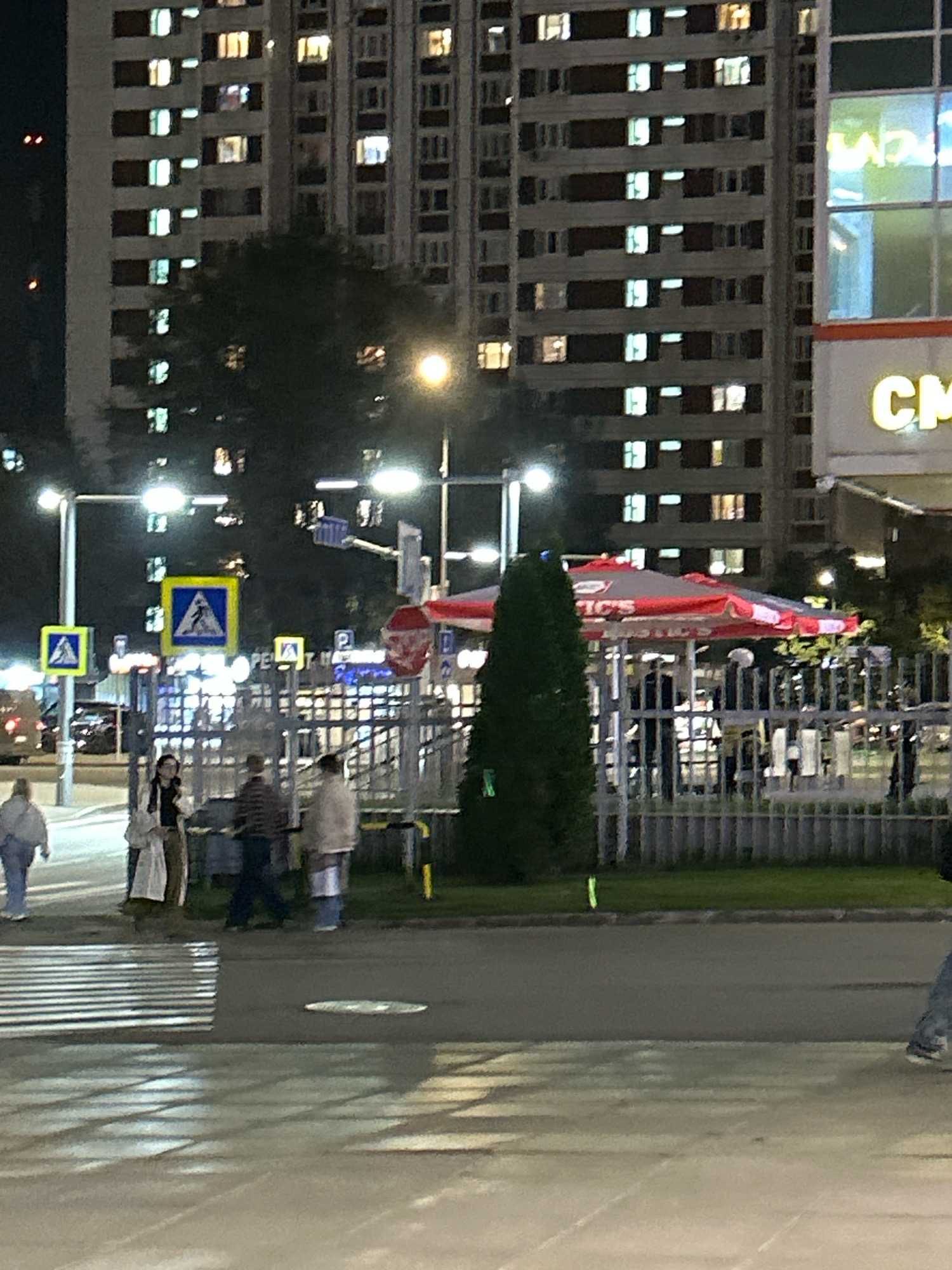 The image depicts a street scene at night in Moscow, Russia, in front of a large, illuminated apartment building. The building features numerous windows, many of which are lit, suggesting occupancy. In the foreground, a pedestrian crossing is visible with white stripes painted on the asphalt. Several people are walking on the sidewalk and near the crossing, including a person in a light-colored coat and headscarf, and two individuals in darker clothing. To the right, a fenced area contains a red and white striped umbrella, possibly from a cafe or kiosk. Streetlights illuminate the scene, casting reflections on the wet ground. Several traffic signs and poles are also present, including a yellow pedestrian crossing sign with a blue border. The overall impression is of a typical urban evening in a residential area.