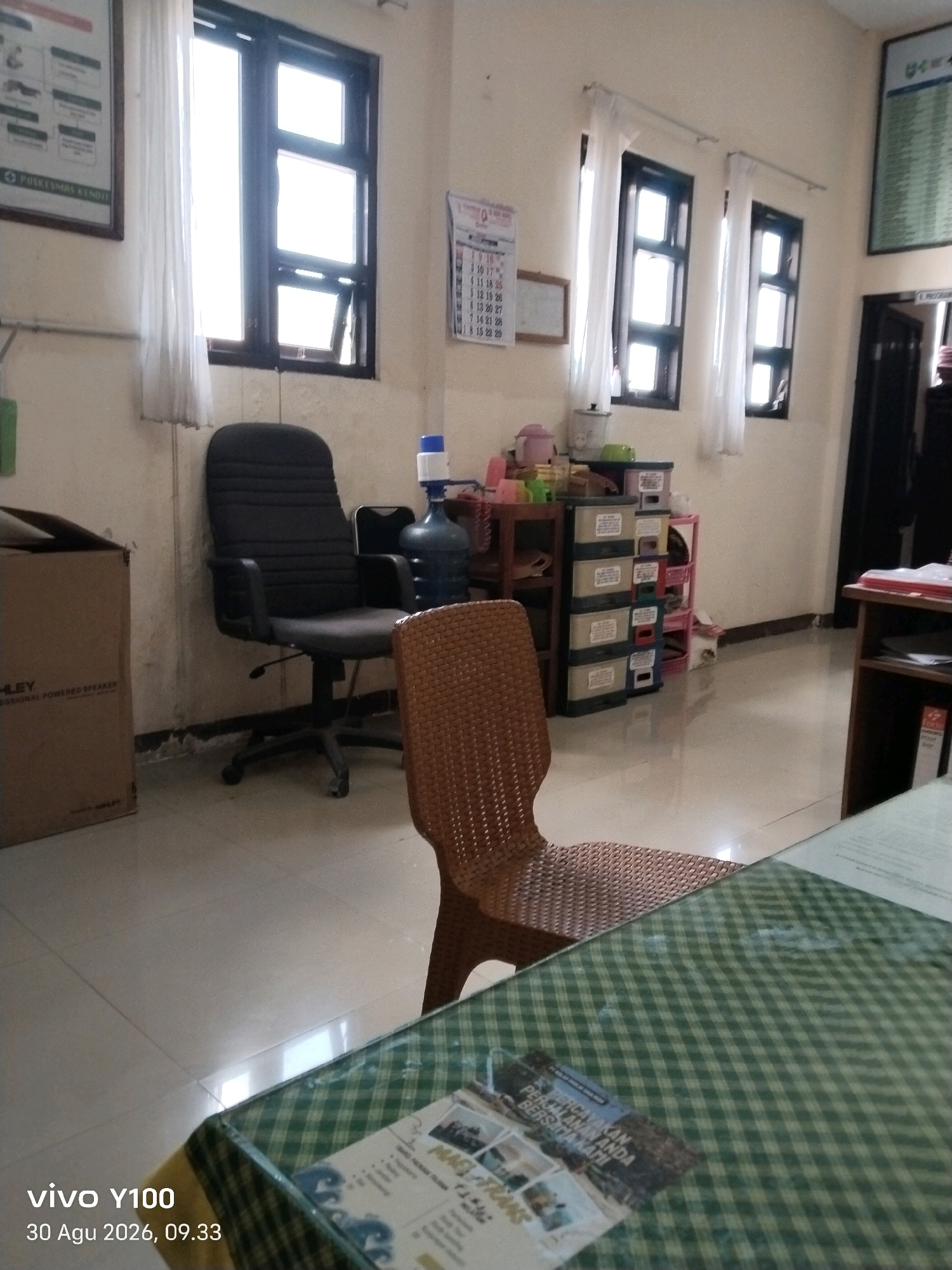 The image depicts an indoor scene, likely an office or administrative space, in Balung, Indonesia. A large, black office chair is positioned centrally in the foreground, angled slightly to the left. To the right of this chair, a smaller, woven brown chair is visible, partially obscuring the office chair. In the background, several large windows with white curtains dominate the wall. To the left of the office chair, a large cardboard box is present, and further left, a bulletin board with text and graphics is mounted on the wall. Adjacent to the windows, a multi-tiered set of drawers is visible, along with a blue water cooler bottle with a white dispenser. The floor appears to be tiled. In the lower right corner of the image, a portion of a table covered with a green and white checkered cloth is visible, with a document or magazine resting on it. The timestamp in the lower left corner indicates the image was taken on August 30, 2026, at 09:33.