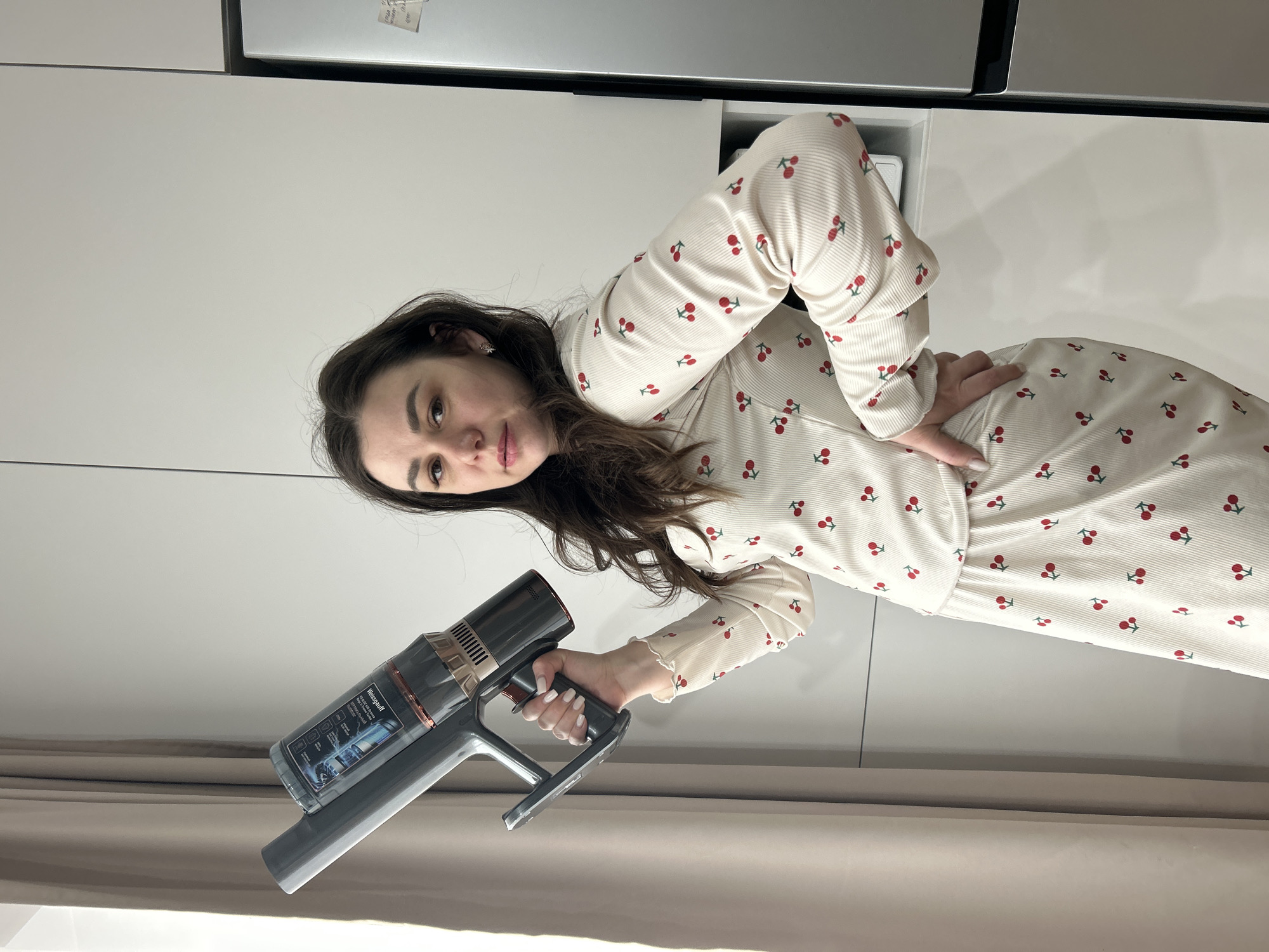 A young woman stands indoors, posing for the camera while holding a handheld vacuum cleaner. She has long, dark brown hair and a neutral expression, looking directly at the viewer. She is wearing a cream-colored, ribbed two-piece outfit (a long-sleeved top and a skirt or dress) adorned with a repeating pattern of red cherries. On her left ear, a small, intricate earring is visible. Her fingernails are painted with white polish.
In her right hand, she holds a dark gray and rose-gold colored handheld vacuum cleaner. Visible text on the vacuum cleaner clearly states "Werngauff". Below this, several features are indicated with icons and small text, including "High-speed digital motor," "Strong suction," "Multi-cone separation," "Long battery life," and "Noise reduction." Her left hand is placed on her hip.
The setting appears to be a modern indoor space, likely a kitchen or utility area, characterized by clean white cabinetry behind her. Above the cabinetry, part of a metallic appliance, possibly a refrigerator or microwave, is visible. The lower part of the image shows beige or light brown curtains, suggesting a window or doorway nearby. The lighting indicates it is daytime, with no specific weather details discernable. The overall atmosphere is casual, portraying a domestic scene. While the location context is Chaikovsky, Russia, there are no specific visual cues within the image to identify this location.