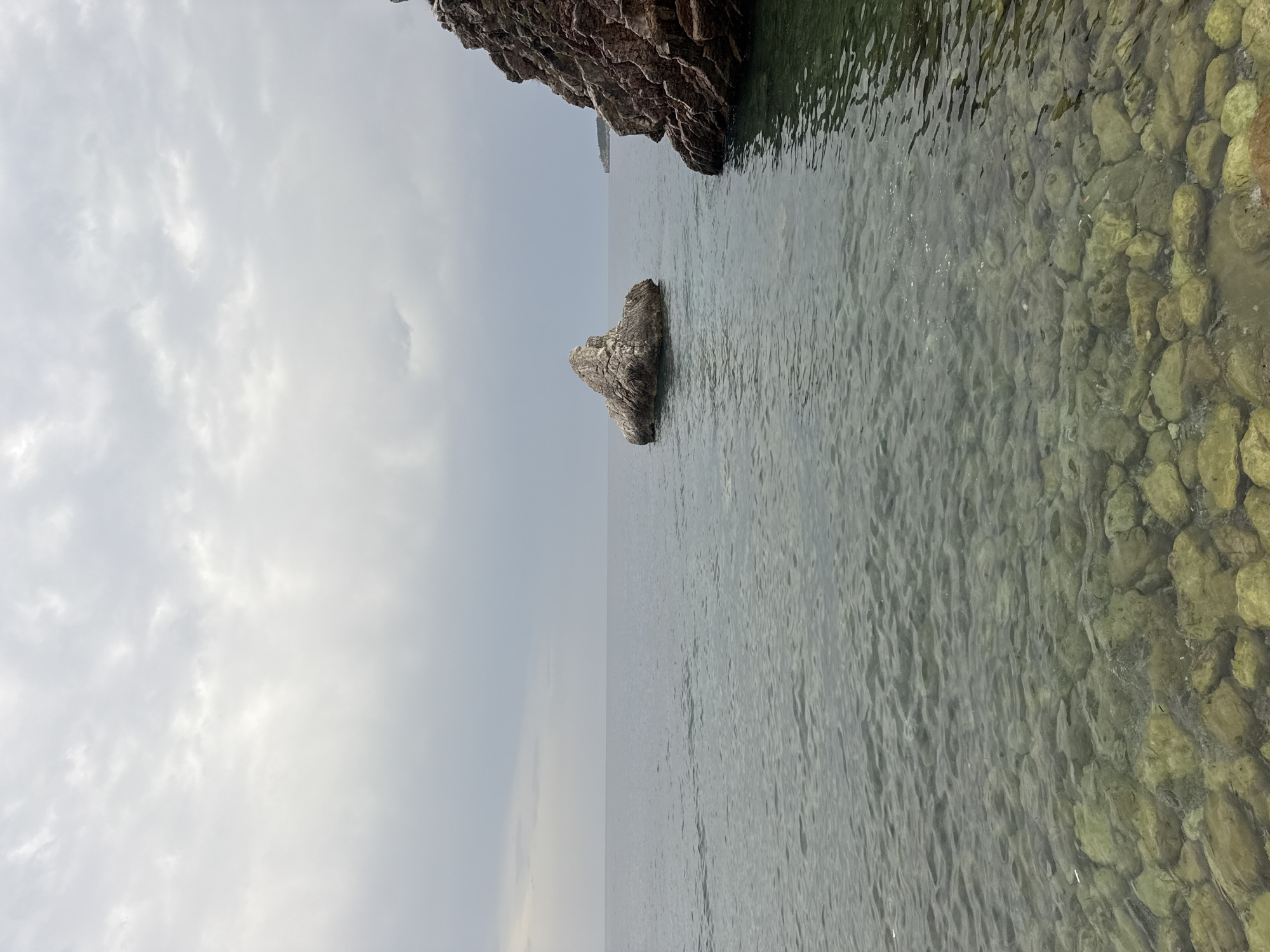 The image depicts a coastal scene in Rafailovići, Montenegro, on an overcast day. The foreground shows a clear, shallow body of water with numerous smooth, rounded rocks visible beneath the surface, indicating a rocky seabed. The water is calm with gentle ripples. In the middle ground, a solitary, large, irregularly shaped rock juts out of the sea. To the right, a steep, rocky cliff face, composed of reddish-brown stone, rises from the water. In the distance, the horizon is marked by a faint line where the sea meets a hazy, overcast sky. The overall atmosphere is somber due to the cloudy conditions.