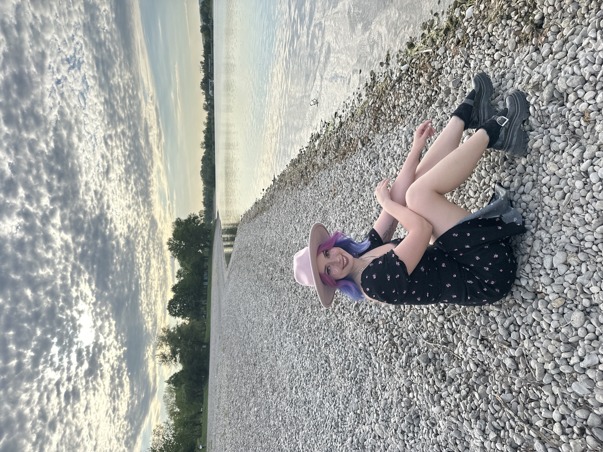 A woman is lying on a pebble beach next to a body of water. She is wearing a black floral dress, black socks, and black shoes with buckles. She has a light pink hat and blue-purple hair. She is smiling and has small, light-colored shapes on her face. The background includes water, a cloudy sky, and trees lining the shore. The image appears to be taken at the City of Zagreb, Croatia.