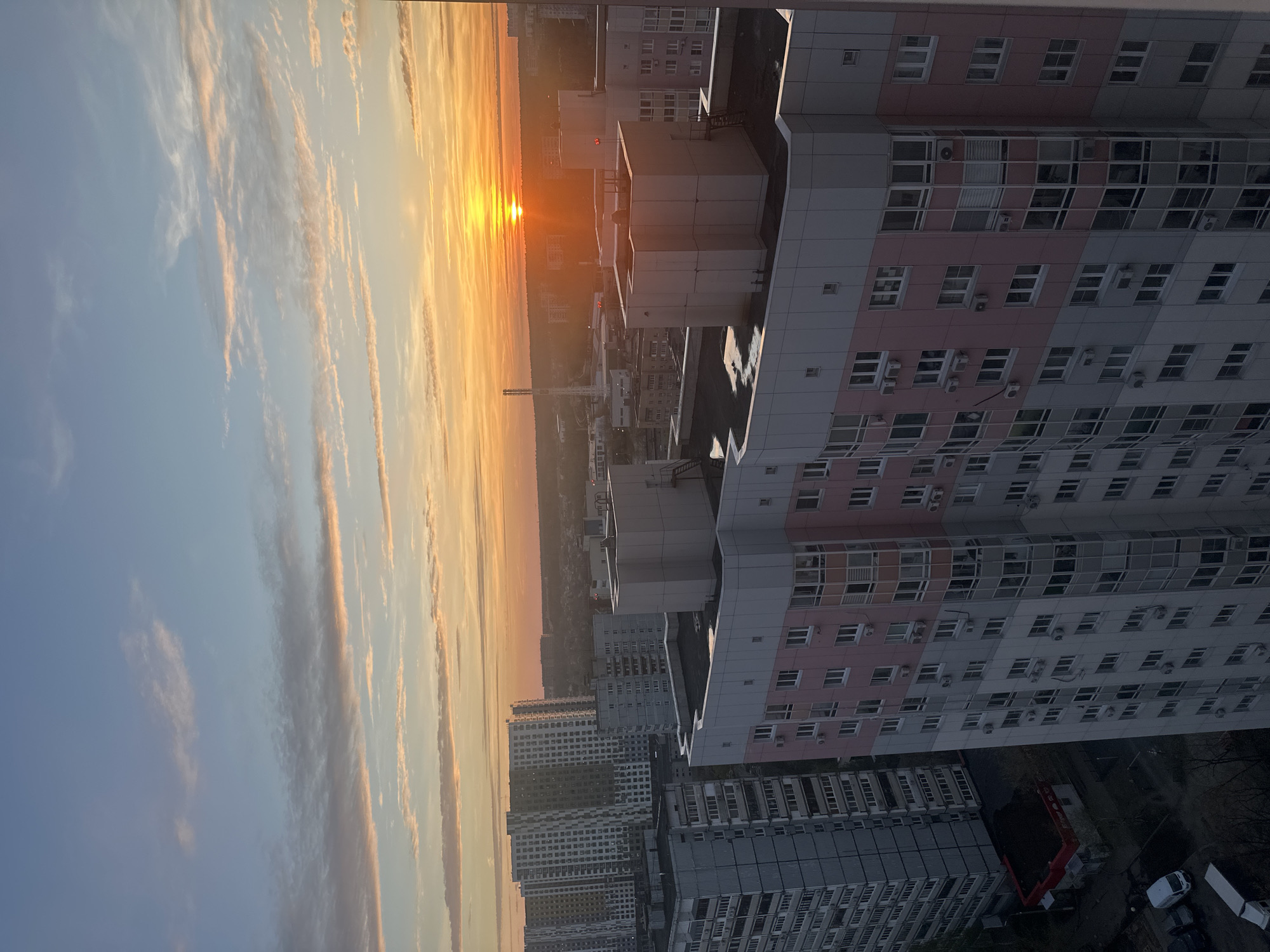 The image shows an urban sunrise in Krasnogorsk, Russia. The sun is rising on the horizon, casting a warm orange glow across the sky and cityscape. The sky is a gradient of blues and oranges, with wispy clouds illuminated by the sunrise. Several high-rise apartment buildings dominate the foreground, their facades a mix of grey, white, and pink. Many windows are visible, some with air conditioning units. In the distance, more buildings and possibly trees can be seen, though details are obscured by the sunrise's glare. The overall scene is a view of a densely populated area during the early morning.
