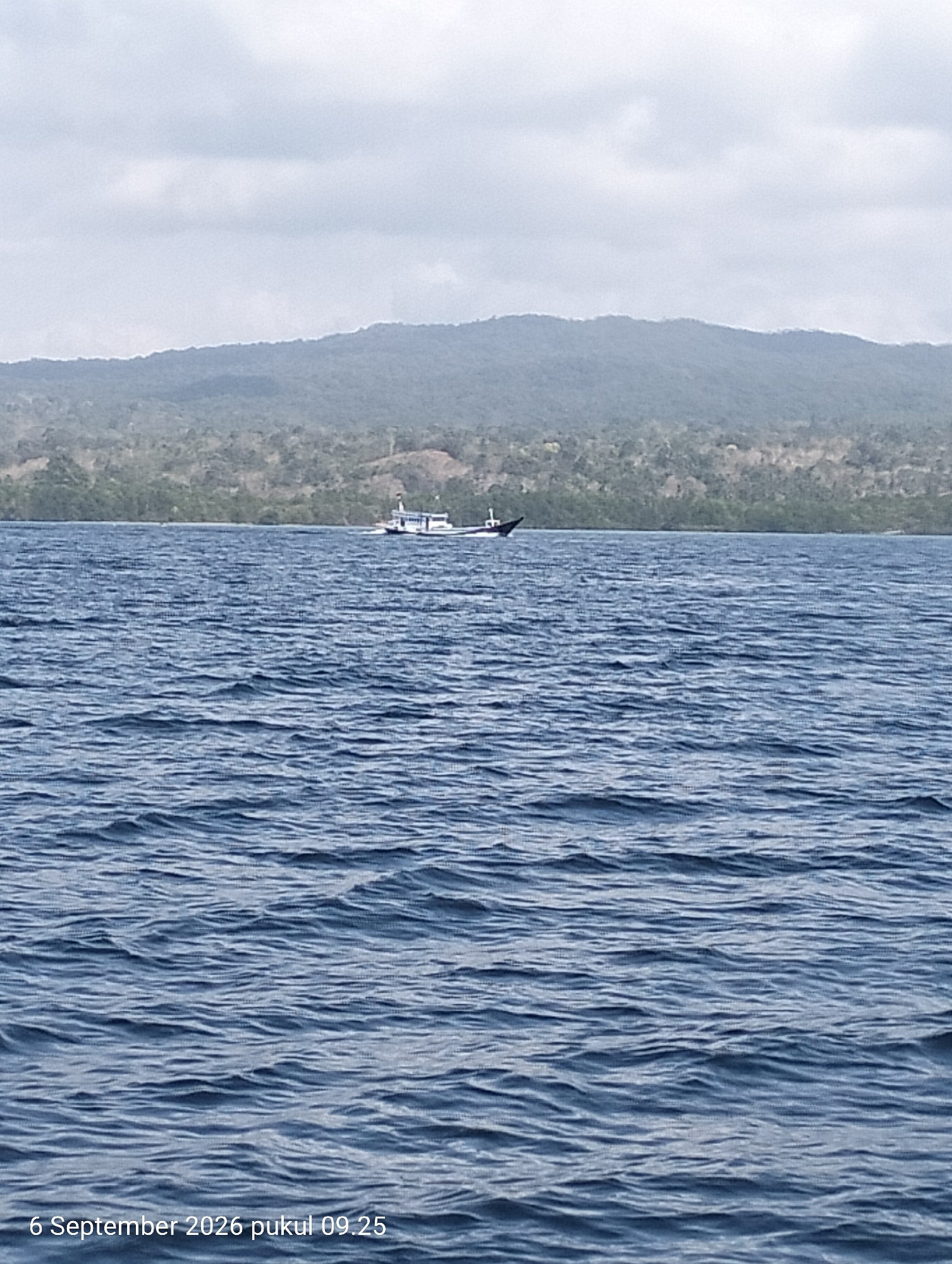 A wide shot captures the vast, choppy blue ocean under a cloudy sky. In the middle distance, a white and blue boat sails across the water. The horizon is marked by a lush, green, tree-covered mountain range. The bottom left of the image displays a timestamp: "6 September 2026 pukul 09.25". The scene is likely along the coast of Kendari, Indonesia, given the timestamp's format and language.