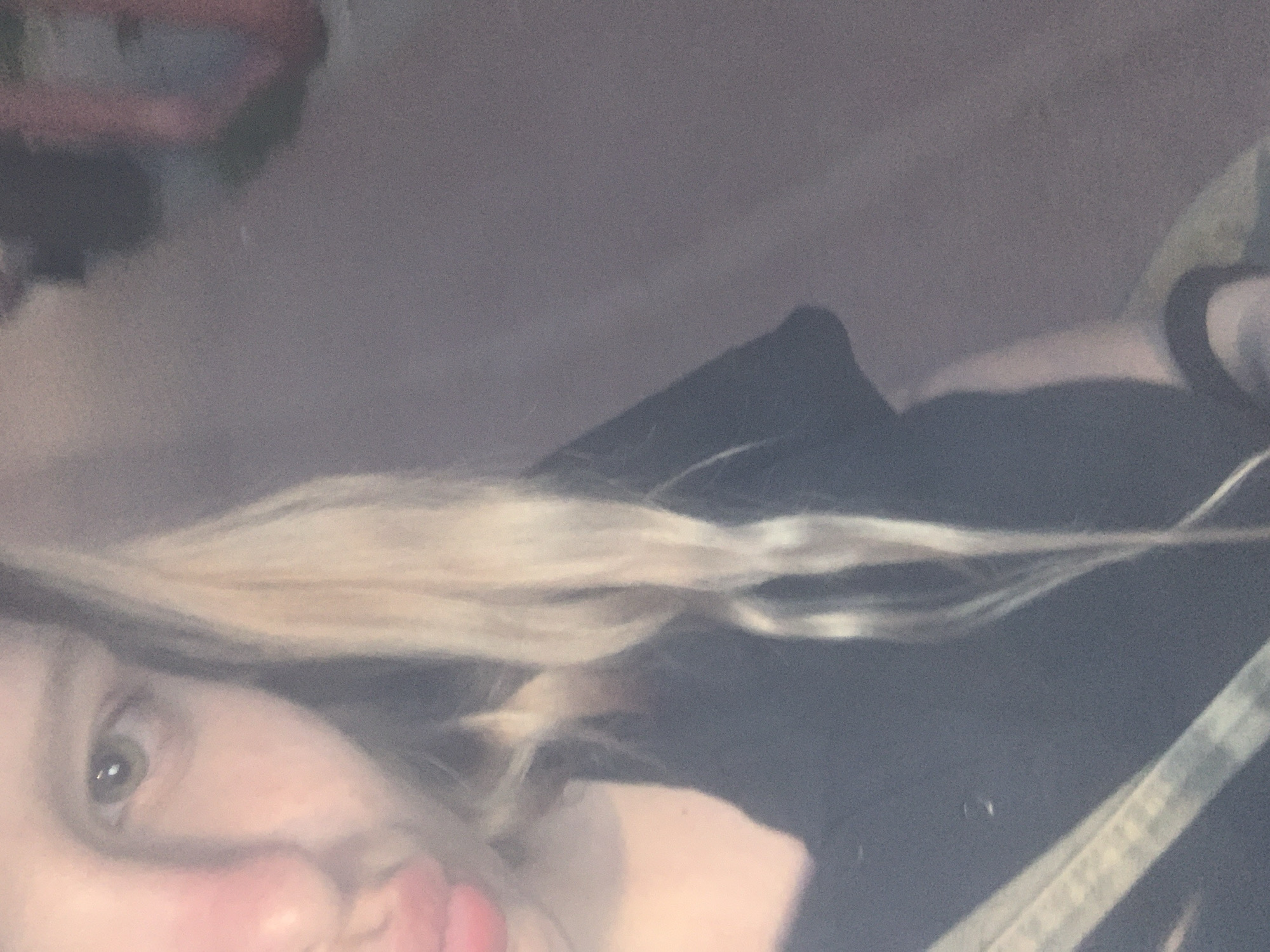 The image shows a close-up, blurry selfie of a person with blonde hair and light-colored eyes. The person is wearing a dark shirt and a strap from a bag is visible across their chest. The background is indistinct and appears to be an indoor setting with muted colors. There is no identifiable information in the image that would suggest a location in Vsevolozhsk, Russia.