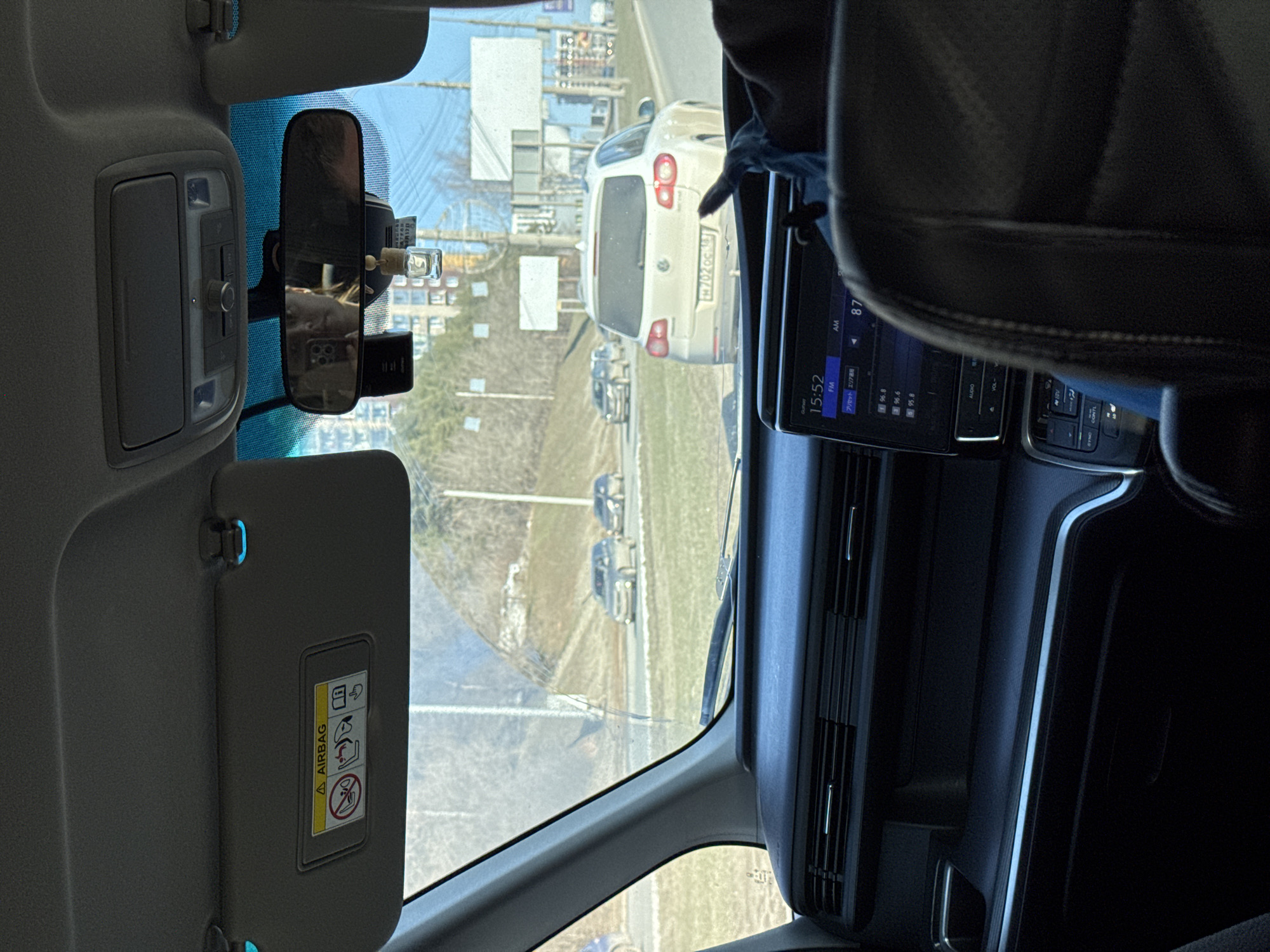 Captured from inside a car, the image offers a glimpse of the outside world through the windshield. The vehicle appears to be stationary on a road with other cars parked and moving in the same direction. Visible through the windshield, we see an off-white Volkswagen Tiguan (license plate p702oc 163). The location is suggested to be in Izhevsk, Russia. Inside the car, we see elements such as the rearview mirror, sun visor, a digital display showing 15:52, and parts of the dashboard and a car seat.