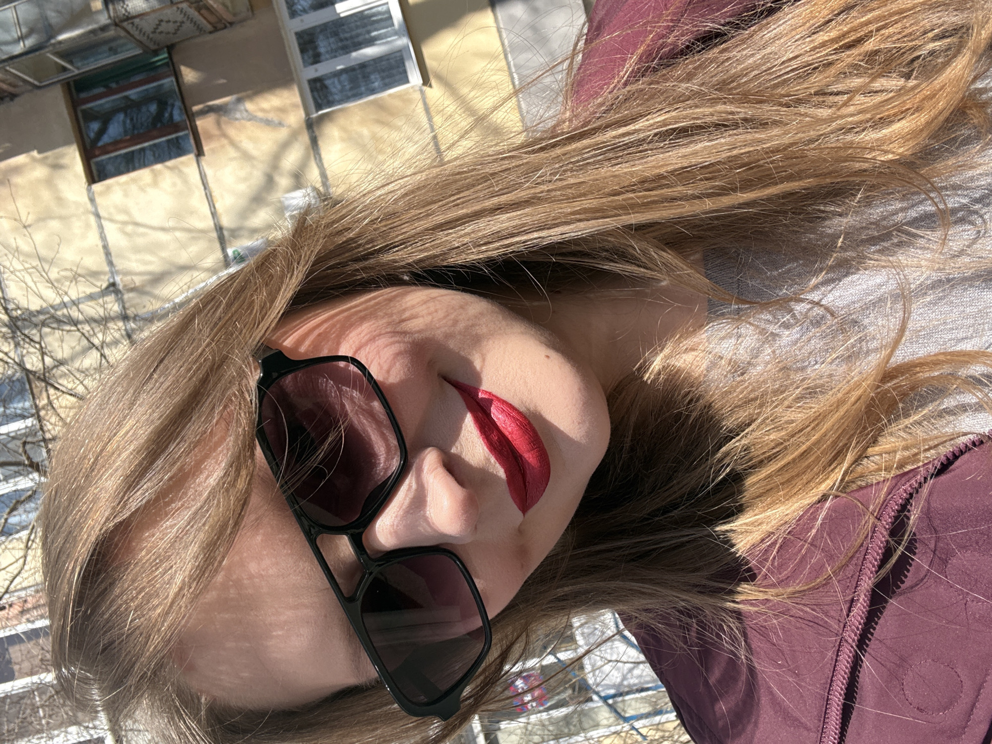 The image presents a close-up, angled portrait of a person, likely a woman, taken outdoors in Izhevsk, Russia. The subject's head is tilted, showing her from mid-forehead down to the collarbone area. She has long, light brown to blonde hair, which frames her face and flows over her shoulder. Her eyes are concealed by large, black-framed sunglasses, and she is wearing vibrant red lipstick. A burgundy or maroon jacket, featuring a visible zipper and subtle circular stitched patterns, covers her upper body.
The background, although blurred, reveals light-colored urban building facades with multiple windows, some reflecting the bright sunlight. Bare tree branches are visible against the buildings and sky, suggesting a cold season like late winter or early spring. The strong, direct sunlight indicates a clear, daytime setting, creating sharp shadows on the subject's face, hair, and jacket, as well as across the background elements. The photograph has an extreme tilt, suggesting a unique perspective, possibly a selfie taken from a low angle or while reclining. No visible text is present in the image.