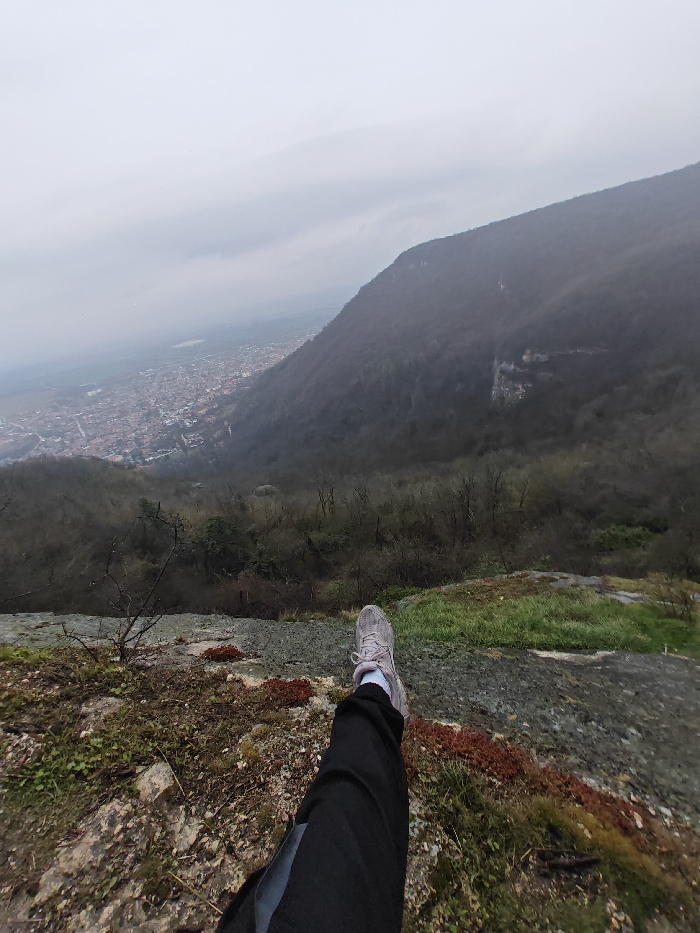 The scene is a first-person, high-angle view from a rocky precipice on the Shumen Plateau, overlooking the city of Shumen, Bulgaria. In the lower right foreground, the lower left leg and foot of an individual are visible, extended horizontally into the frame. The individual wears black trousers, a light-colored sock, and a grey athletic shoe with white laces. The immediate ground around the foot consists of rugged rock formations interspersed with patches of green moss, reddish-brown vegetation, and sparse dry plant matter.
Below the observer, a steep, densely forested hillside descends into the valley, covered predominantly by bare deciduous trees and exhibiting several exposed rock outcrops or small cliff faces. In the background, the city of Shumen is spread across the valley floor, characterized by a dense arrangement of buildings, many featuring red-tiled roofs. Beyond the city, a body of water, possibly a reservoir, is discernible in the far distance. The sky is uniformly overcast with a light grey cloud cover, and atmospheric haze reduces clarity of distant features.