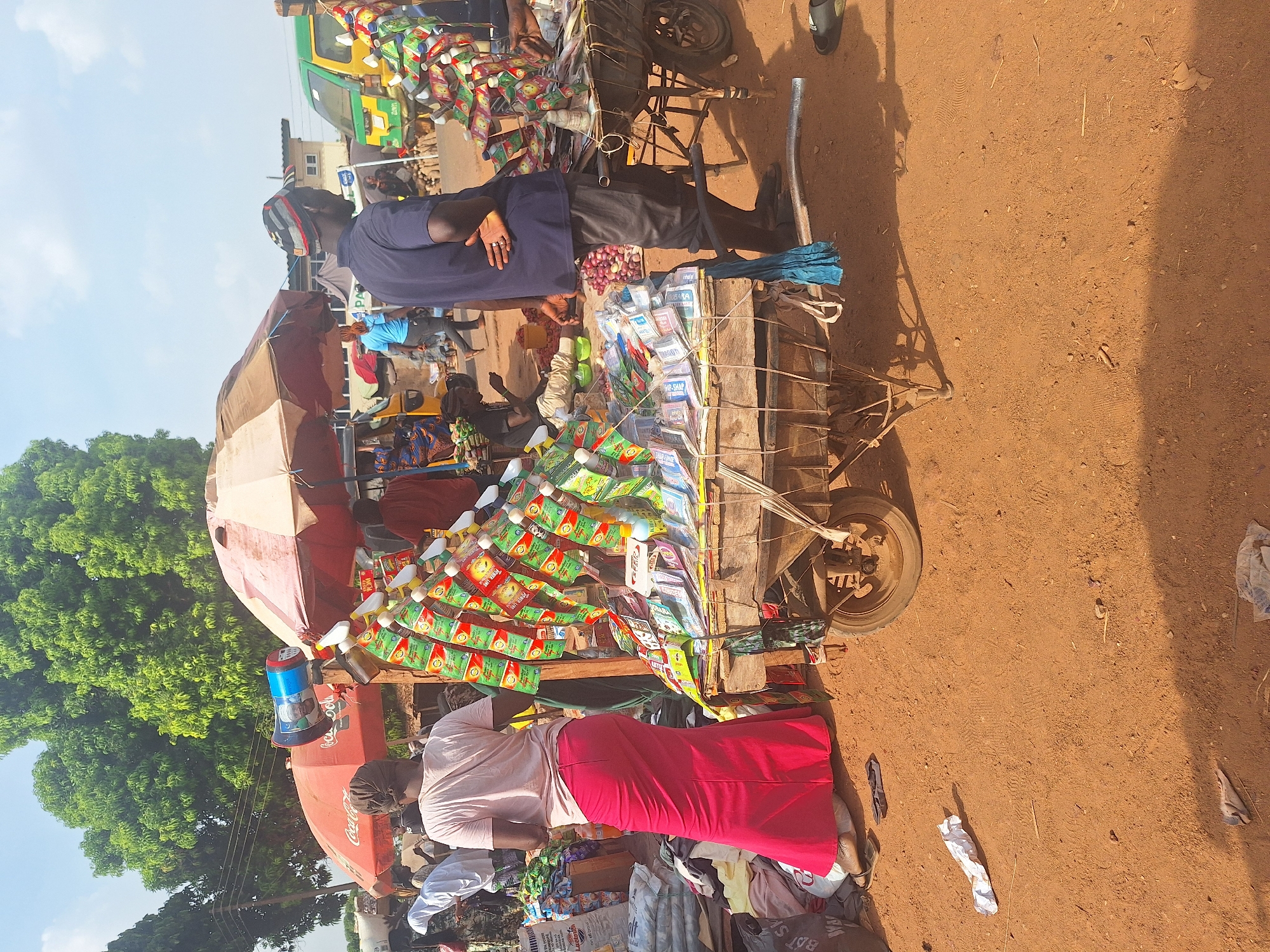 The image shows a street scene in Kakuri Gwari, Nigeria. A mobile vendor is operating from a three-wheeled cart, selling a variety of packaged goods, including condiments and household items. The cart is laden with merchandise and is partially covered by a Coca-Cola branded umbrella. The ground is reddish-brown, suggesting the soil composition in the area. Several individuals are present, including the vendor and at least one customer, a woman in a pink skirt. A man in a blue shirt stands near the cart. A smaller hand cart is visible in the background.
