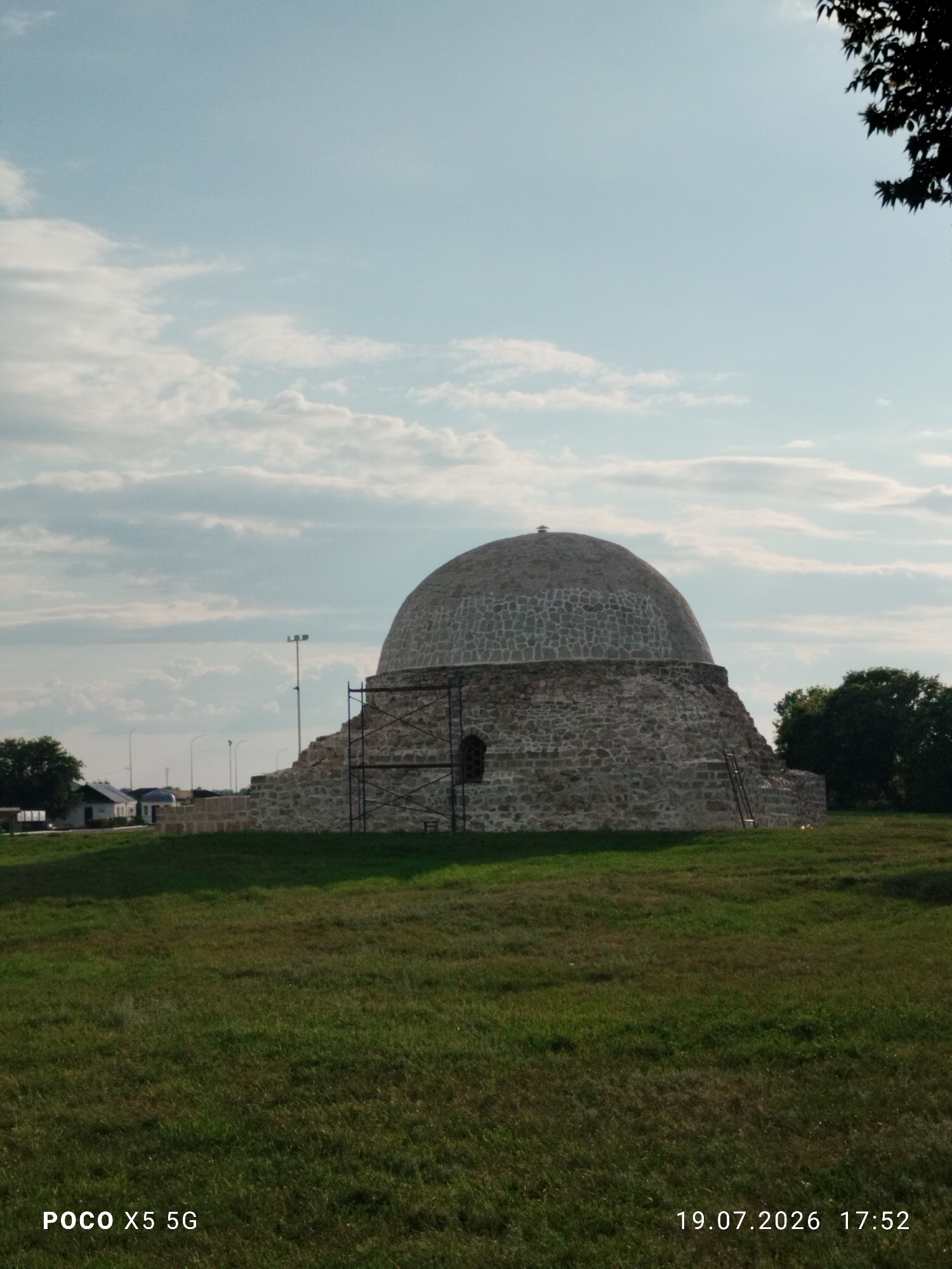 The image shows a view of the Black Chamber in the city of Bolgar, Russia. The Black Chamber is a historical monument, a domed building constructed of stone. It is surrounded by a grassy field under a cloudy sky. In the foreground, there is a ladder leaning against the structure and scaffolding around a portion of the building. In the background, there are some smaller white buildings and trees. The lower part of the image shows the text "POCO X5 5G" and the date "19.07.2026 17:52".