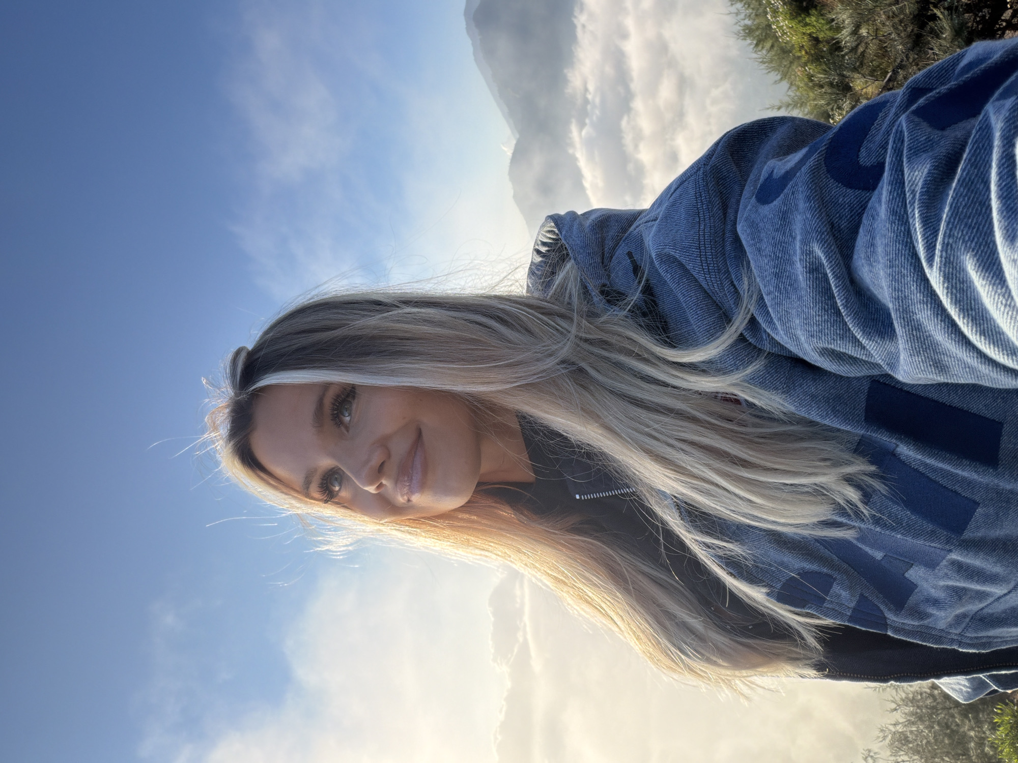 A young Caucasian woman with blonde hair is captured in a selfie taken from a low angle. She is wearing a blue denim jacket with dark blue accents and a black hoodie underneath. Her head is tilted back as she looks up towards the sky, and she has a gentle smile on her face. The background reveals a breathtaking view from Kintamani, Indonesia. Below her, a sea of clouds blankets the landscape, with the misty peaks of mountains visible through the ethereal fog. The sky above is a clear blue, with a few wispy clouds and the soft glow of sunlight. The scene conveys a sense of peace and wonder, as if the woman is taking in the majestic beauty of the early morning or late afternoon.