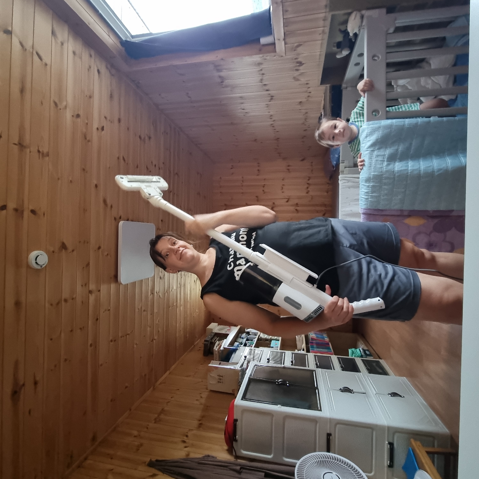In Dunnae-myeon, South Korea, a woman stands in a room with wood-paneled walls and ceiling, holding a white cordless vacuum cleaner. She wears a black tank top with white text and gray shorts. In the background, a young child in a green and white striped shirt is visible in a crib, partially covered by a blue blanket. To the left of the woman, a white fan and a stack of white plastic drawers are present. Shelving units filled with books and various items are visible behind the drawers. A window with a dark curtain is on the right side of the room. The overall environment appears to be a residential indoor space.