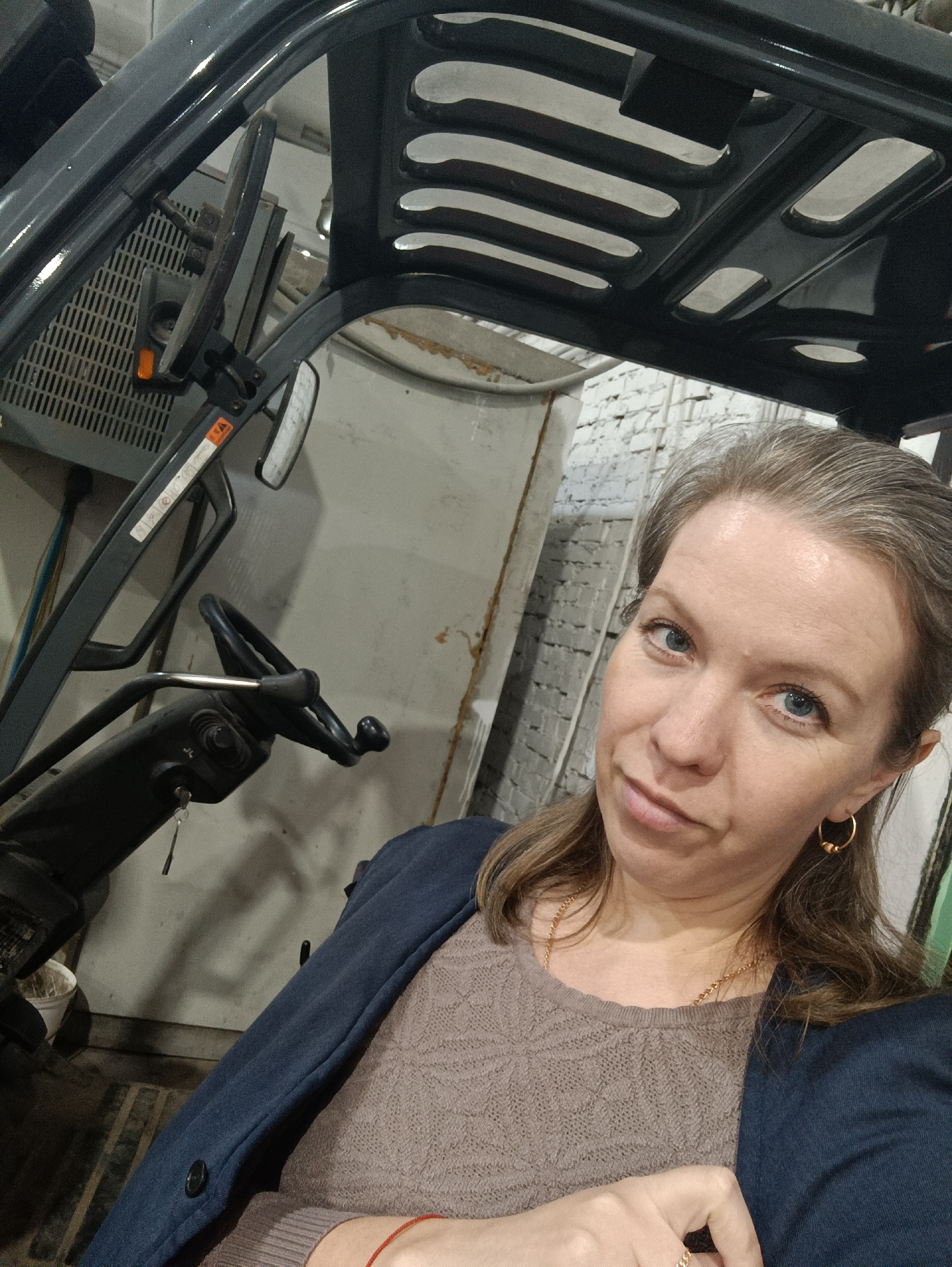 A woman is seated inside what appears to be a forklift or similar industrial vehicle. She has fair skin, blue eyes, and light brown hair with some graying. She is wearing gold hoop earrings, a brown sweater with a textured pattern, a blue blazer, and a gold necklace. She is looking directly at the camera with a slight smirk.
The vehicle's interior is visible, including the steering wheel, dashboard, and protective overhead structure. The vehicle seems to be parked indoors, possibly in a warehouse or industrial space. The background includes a partially visible white wall, some equipment, and indistinct items.
There is no information about the location of the city of Chaikovsky, Russia in the image, so the city is not part of the scene.