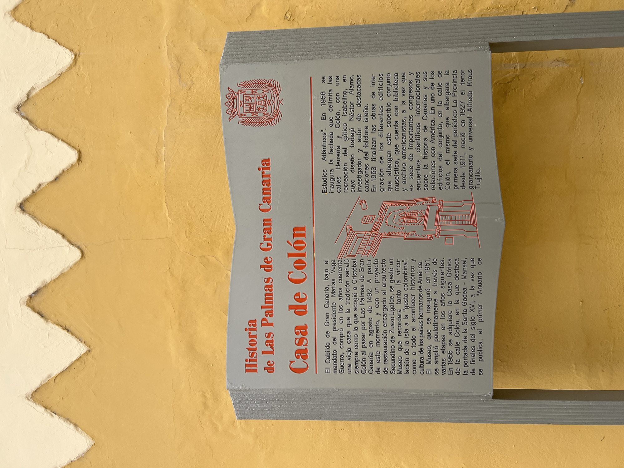 The image shows a placard in Las Palmas de Gran Canaria, Spain, detailing the history of the Casa de Colón (Columbus House). The sign features red lettering against a white background. The text explains the origins of the house as a place where Christopher Columbus stayed on his way to the Americas in 1492. It describes its transformation into a museum dedicated to the history and culture of the Americas and its relationship with the Canary Islands. The text highlights the architectural aspects, including the incorporation of the Casa Gótica and the gótico isabelino style facade. The environment is a wall.