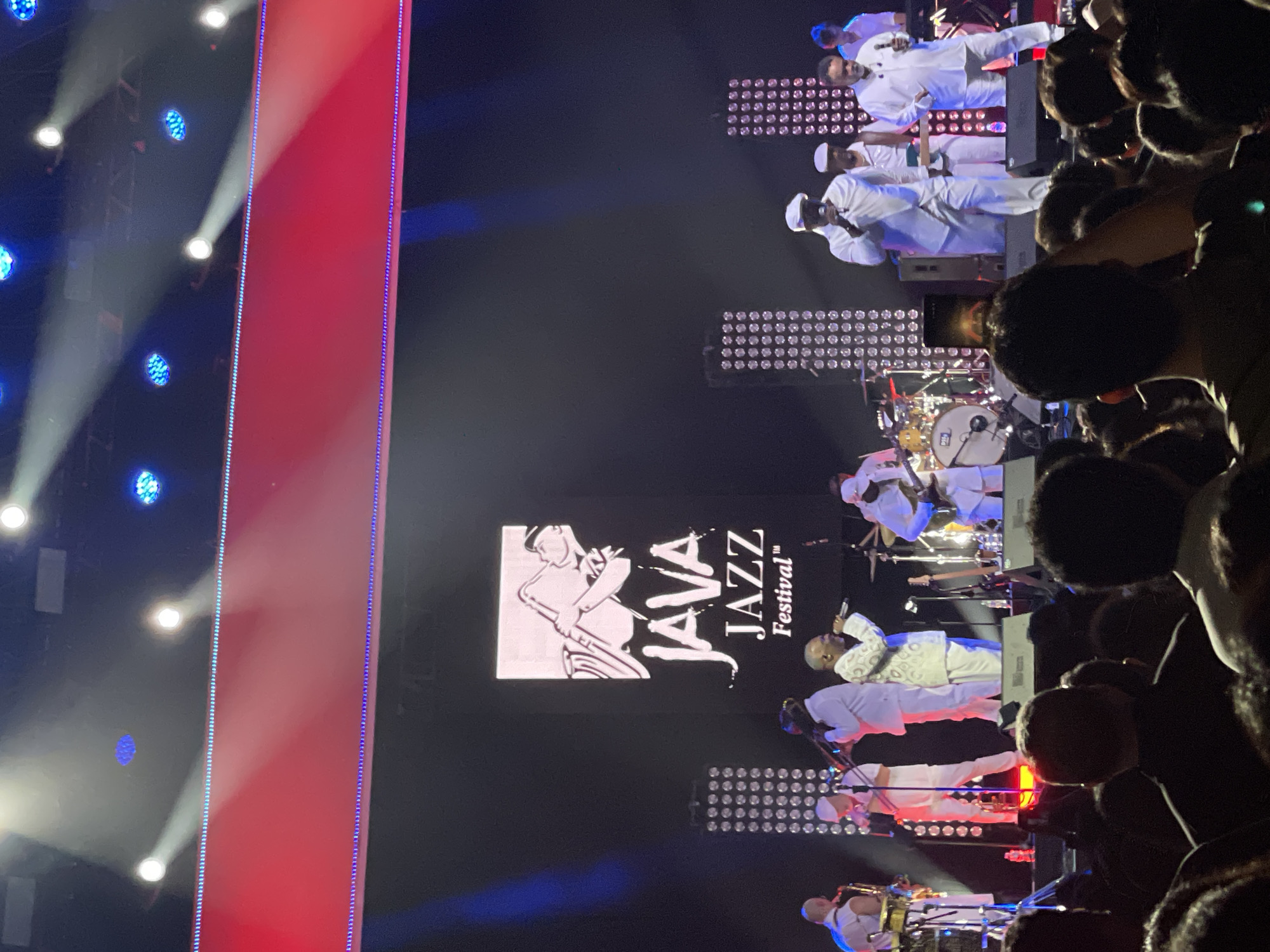 The image shows a live music performance at the Java Jazz Festival in Salembaran, Indonesia. The audience is visible on the right, with heads and shoulders of people densely packed. On the stage, a band dressed in white uniforms is performing. A screen behind the performers displays the Java Jazz Festival logo. There are stage lights above and behind the band. A large red light panel is seen on the left side of the stage.