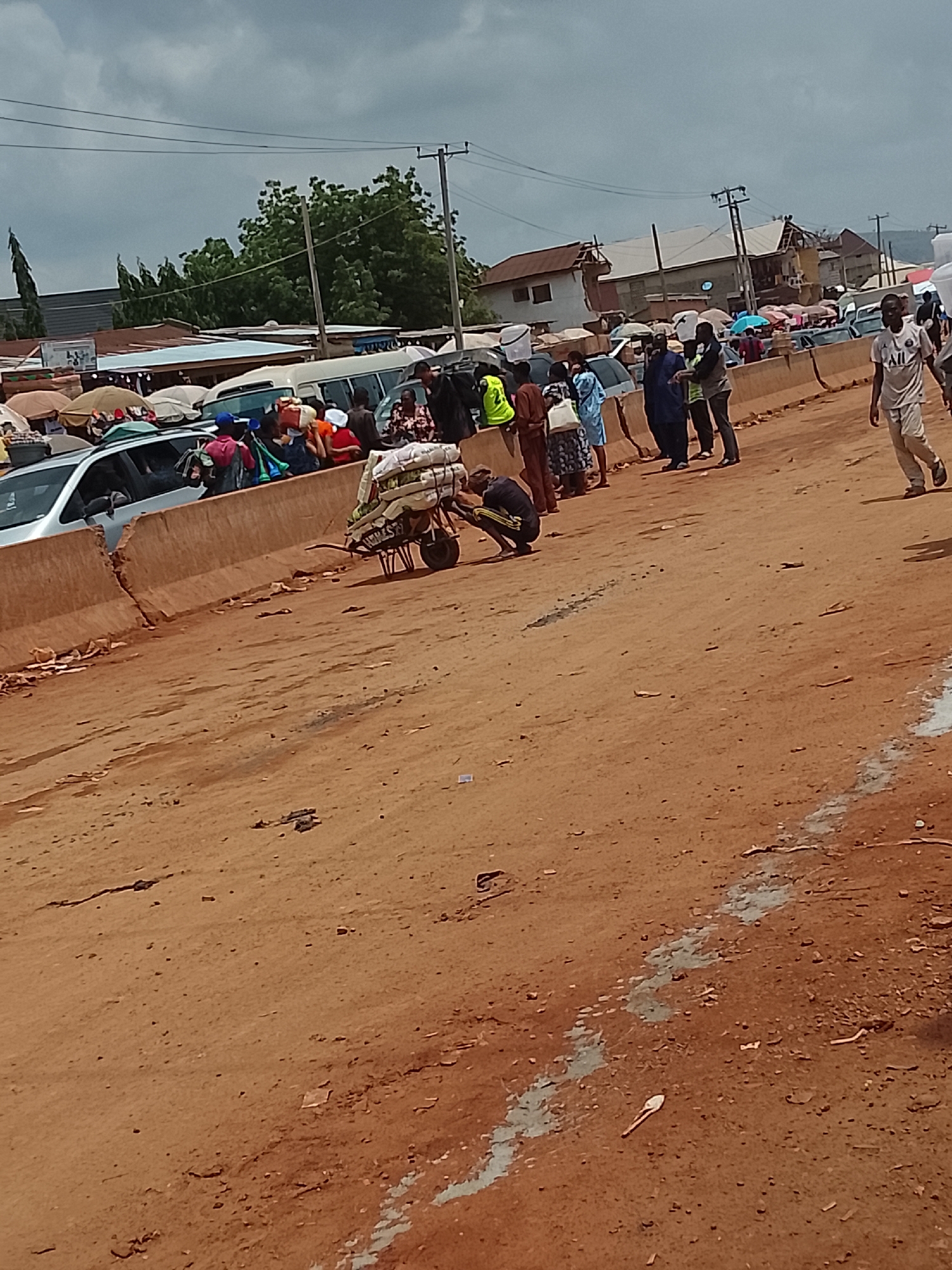 The image depicts a bustling market scene in Katambe, Nigeria. The ground is reddish-brown, appearing to be a dirt road or unpaved area. A concrete median divides the scene horizontally, with people gathered on both sides. On one side, vendors are positioned behind parked vehicles and under umbrellas, likely selling goods. People are clustered in groups, some standing and some moving about. A wheelbarrow loaded with goods is visible in the foreground. A man in a white shirt walks along the edge of the scene. The background consists of buildings and trees, with overcast skies above.