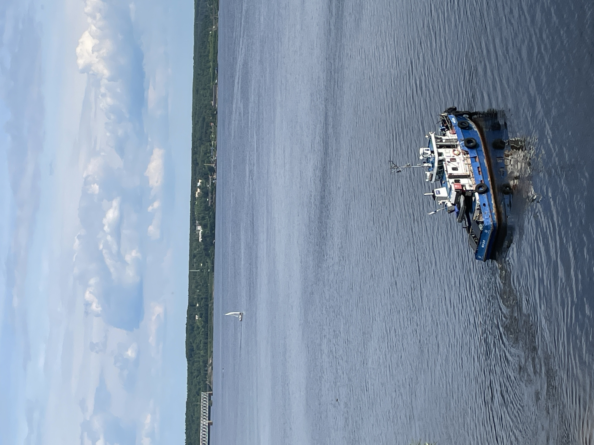 The image captures a wide expanse of water, likely a river, under a bright, partly cloudy sky. In the foreground to the right, a blue and white boat, resembling a tugboat or workboat, is prominently visible. It appears to be listing significantly or partially submerged, with its stern notably lower in the water, creating a pattern of ripples and disturbed water around it. Several black tires are visible hanging along its side, serving as fenders. Faint text, possibly "8160", is discernible on the stern of this blue boat.
Further in the distance, a smaller white sailboat with a single sail is seen gracefully gliding on the water. The far bank of the river is lined with a continuous stretch of lush green trees, indicating a forested area, interspersed with a few scattered buildings or structures. On the left side of the frame, close to the distant bank, a section of what appears to be a pier or a bridge structure is visible. The scene depicts a calm daytime setting, likely late morning or afternoon, in Пятый километр, Russia, with the listing blue boat being the central point of interest.