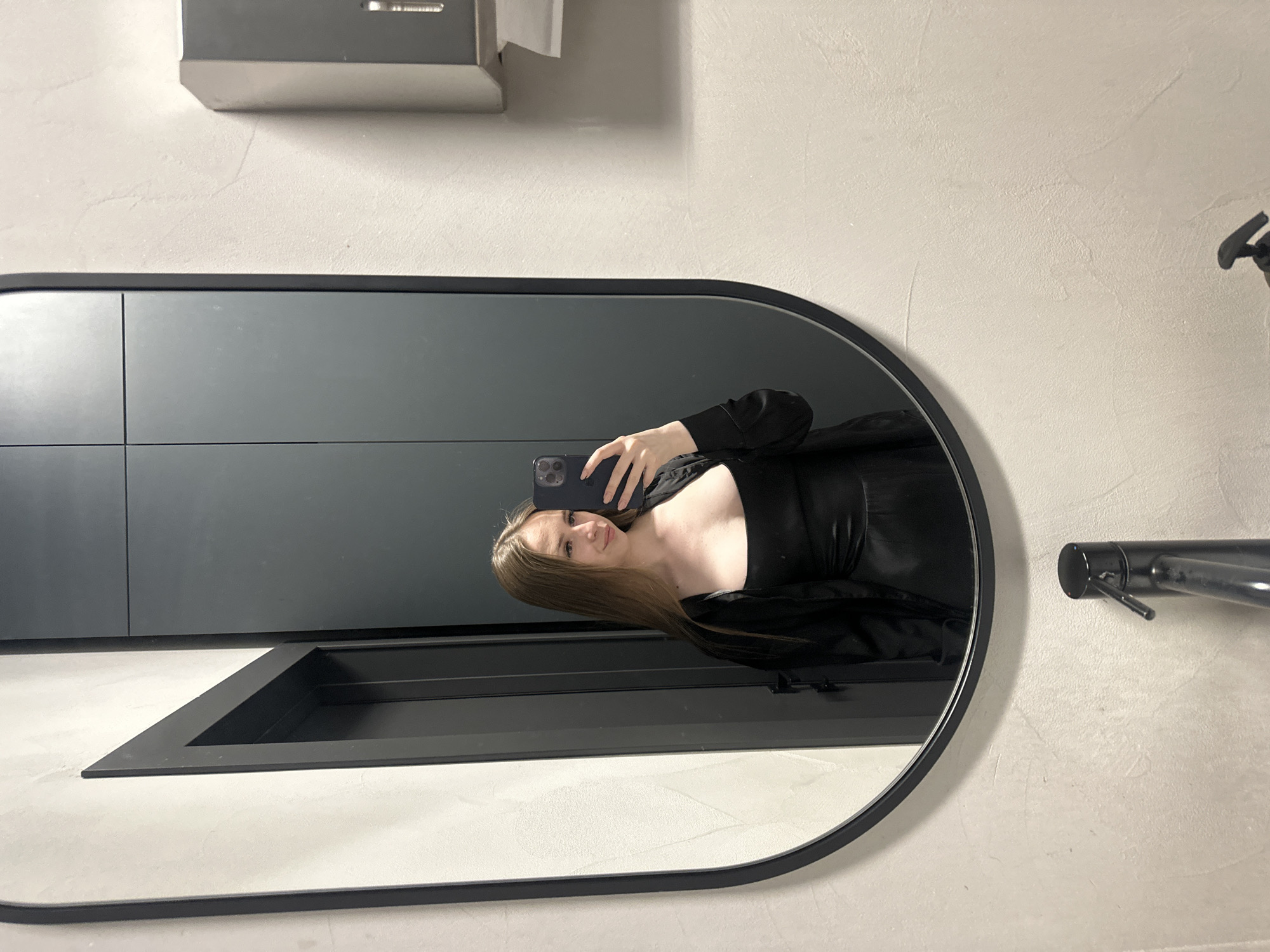 A young woman with long, brown hair stands in front of a large, oval mirror, taking a selfie. She is wearing a black dress with a square neckline and a black jacket. The mirror reflects the woman, as well as a gray door behind her and a wall with a textured finish. To the right of the mirror, a black faucet is visible on a sink. A gray paper towel dispenser is mounted on the wall above and to the right of the mirror. The scene appears to be in a bathroom, possibly in a public or commercial establishment. The lighting is bright and even.