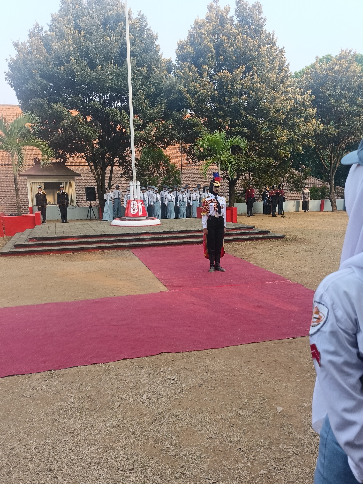 This image depicts an outdoor flag-raising ceremony taking place in front of a brick building, likely in the city of Pengkol, Indonesia. In the foreground, a person in a distinctive white and red uniform with a feathered headdress stands on a red carpet, facing away from the camera. Behind them, a group of students in school uniforms are assembled. A flagpole with a white flag stands prominently in the center of the scene, next to a large red structure with the number "81" displayed on it. Several adults are also visible in the background, some standing and others speaking into a microphone. The ground is a mix of dirt and dry grass, with a large red carpet extending from the foreground towards the ceremony area. Palm trees and other foliage surround the building and the open space. The sky is overcast, suggesting it may be early morning or late afternoon.