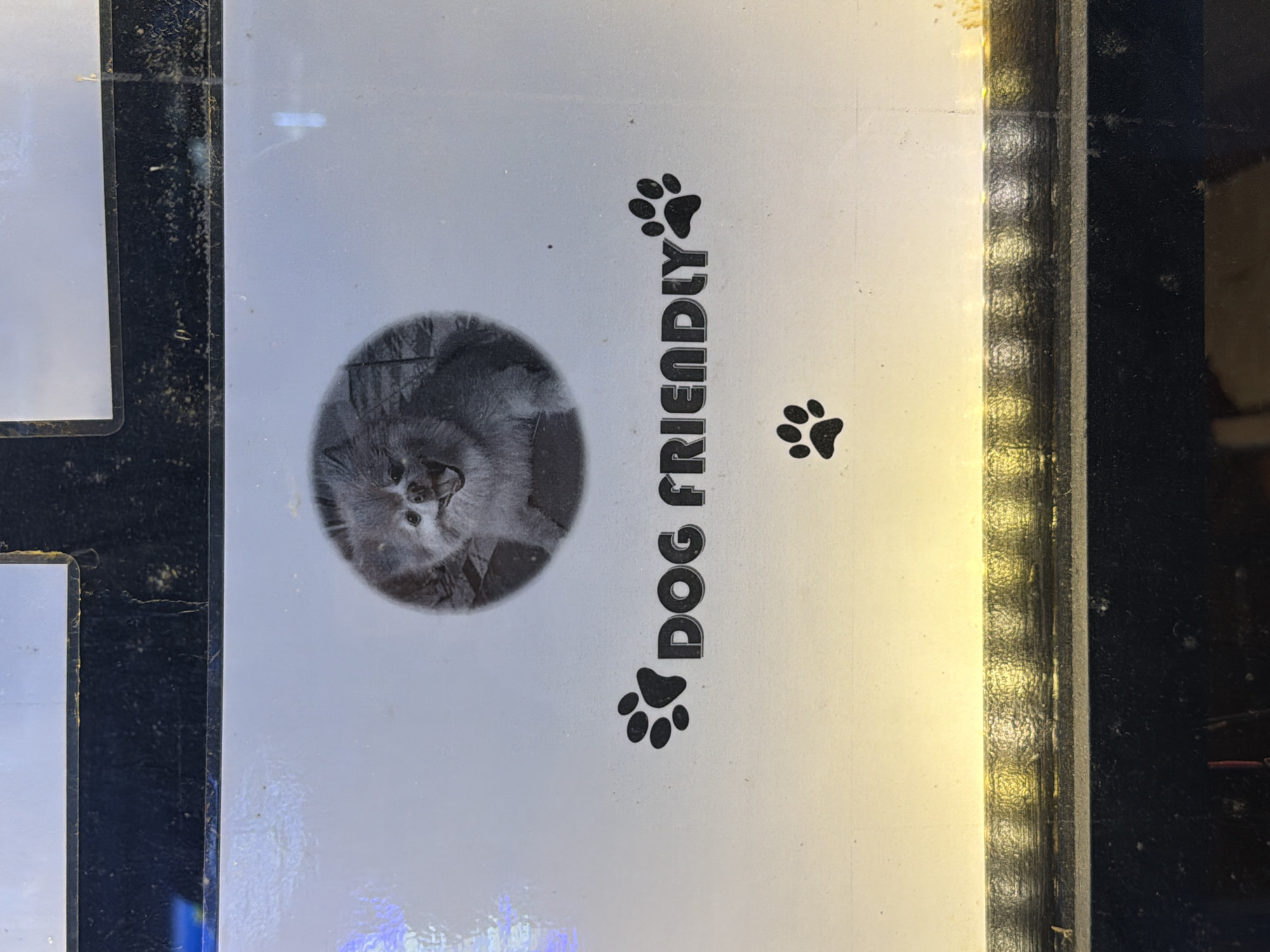 The image is of a "Dog Friendly" sign. It features a gray, fluffy dog in a circular frame, the words "DOG FRIENDLY" in a vertical column with paw prints above and below, and a smaller paw print near the top. The sign is attached to a black frame, which is dirty. The overall lighting suggests the sign is indoors, possibly illuminated by a light source to the right. There is no identifiable information to place this image in London, United Kingdom.