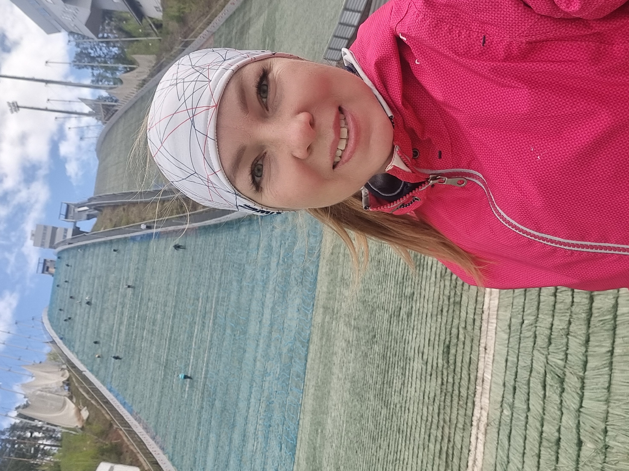 A woman in a pink jacket and white patterned headscarf is smiling and posing for a selfie in front of the ski jumping complex in Прикамский, Russia. The large ski jump is visible behind her, with its artificial turf surface and scaffolding. A few people are walking on the ski jump. The weather appears to be sunny with a few clouds in the sky.