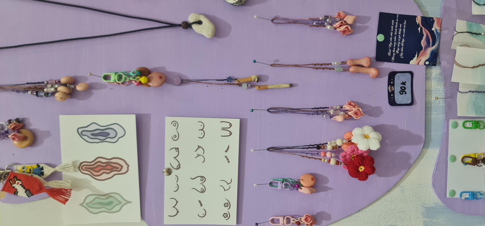 The image shows a display of handmade jewelry and artwork, likely for sale. The background is a light purple surface with various items pinned to it. The items include necklaces, beaded accessories with what appear to be breast-shaped components, crochet flowers, and keychains. There are also cards with drawings, including diagrams of breasts and abstract organic shapes. A price tag displays "90k" (likely 90,000 Vietnamese Dong), suggesting the location is in Vietnam, possibly a market or craft fair. A card includes text in Vietnamese, which translates to a wish for good fortune and happiness. The overall impression is of a stall or shop showcasing handmade crafts, specifically jewelry, with a distinct aesthetic. The city of Đà Nẵng, Vietnam, is not visible in the provided image.