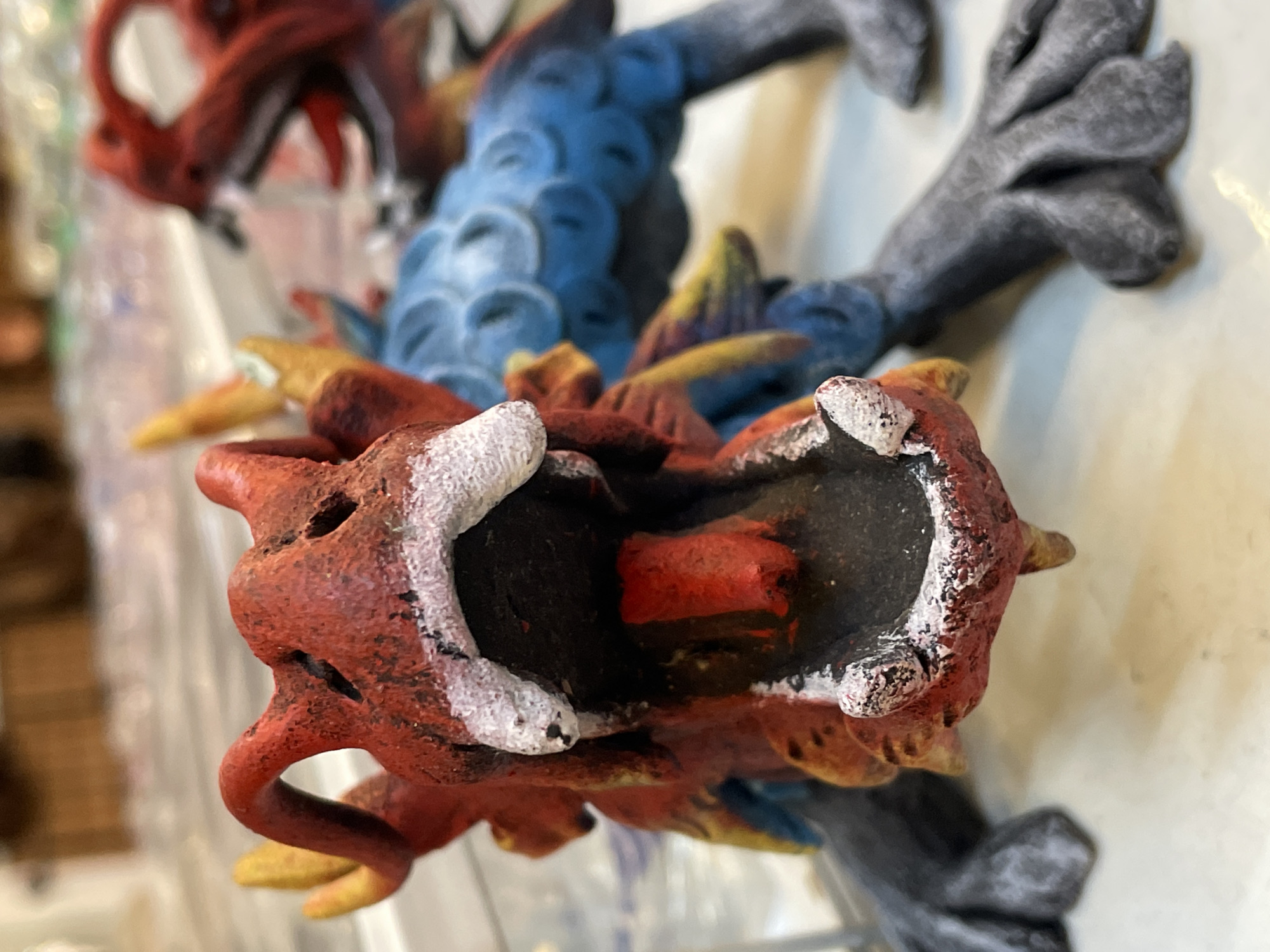 The image shows a close-up of a red and blue dragon-like figurine, possibly a toy or decorative item. The figurine has its mouth open, revealing a red tongue. The red part of the figurine appears textured and painted with black and white accents around the mouth. The blue part has a scale-like texture. The figurine is positioned on a light-colored surface, and its feet are visible, with one foot in the foreground appearing to have four toes. In the background, there is a blurred view of what could be shelves with other objects, suggesting an indoor setting such as a shop or a display. The lighting suggests it's an indoor environment with diffused light. No specific location in Pattaya City, Thailand can be identified from the image.