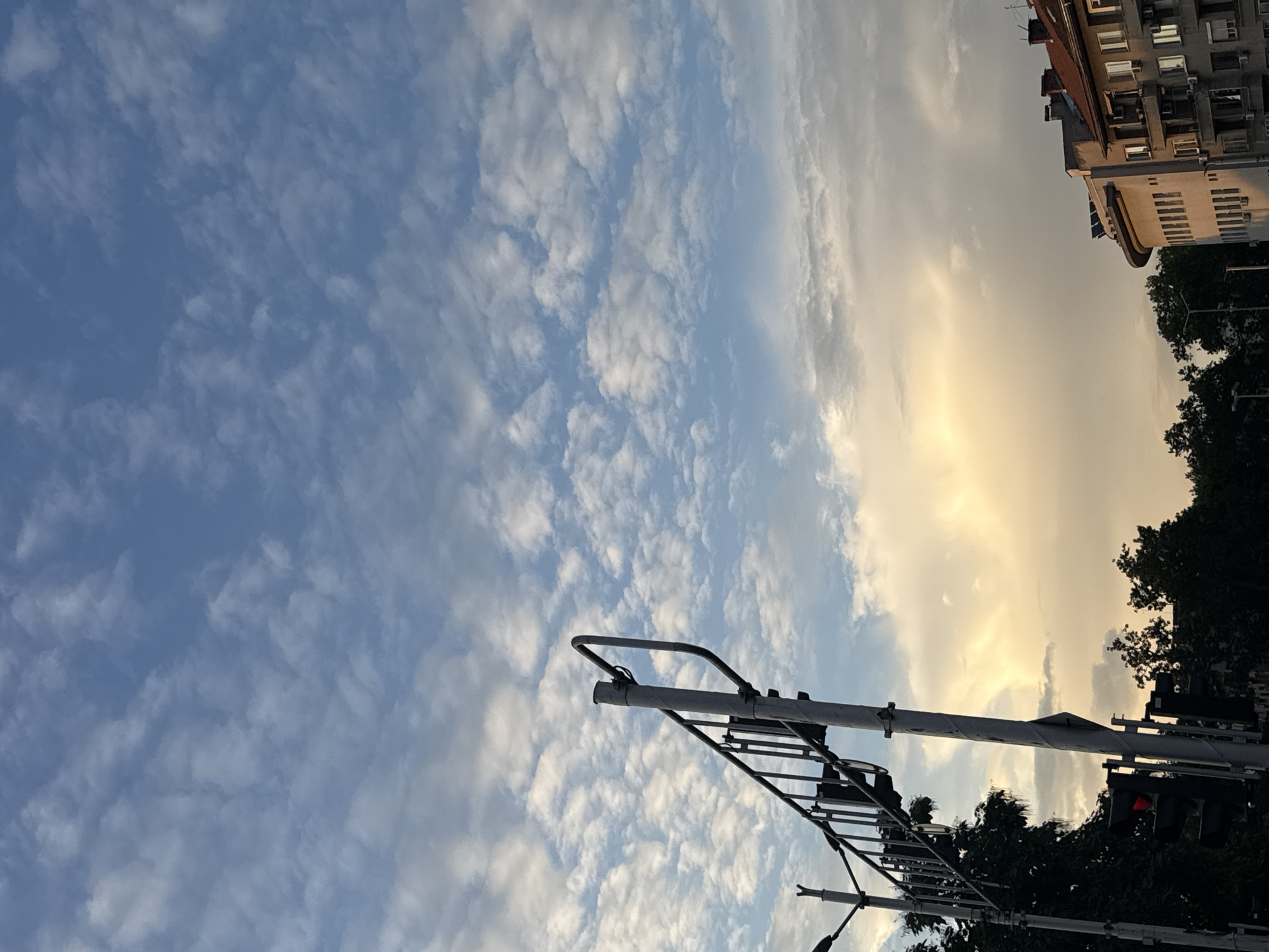 This is a photograph of a street scene in Plovdiv, Bulgaria, taken from a low angle looking up at the sky. The sky is a mix of blue and golden clouds, suggesting it is either sunrise or sunset. A traffic light with a red light illuminated is visible on the right side of the image, along with the top of a building. There is also a metal pole with what appears to be a ladder attached to it, positioned in the foreground and extending upwards towards the sky. The overall impression is of an urban environment at the beginning or end of the day, with the elements of daily life such as traffic control and buildings juxtaposed against the natural beauty of the sky. There is no visible text in the image.