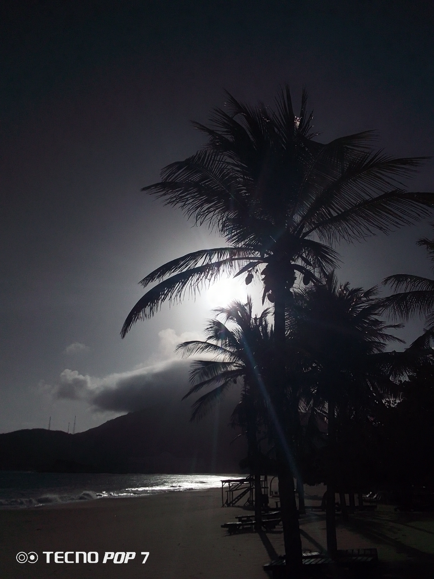This image shows a beach in Pedro González, Venezuela. Palm trees dominate the foreground, silhouetted against a bright, overcast sky. The sun is visible through the trees, creating a lens flare. In the middle ground, a sandy beach leads to the ocean, with waves reflecting the sunlight. In the background, a dark landmass rises, partially obscured by clouds. Beach furniture, including a structure and benches, can be seen along the edge of the beach. The image has a dark, high-contrast appearance. The text "TECNO POP 7" is displayed in the lower-left corner.