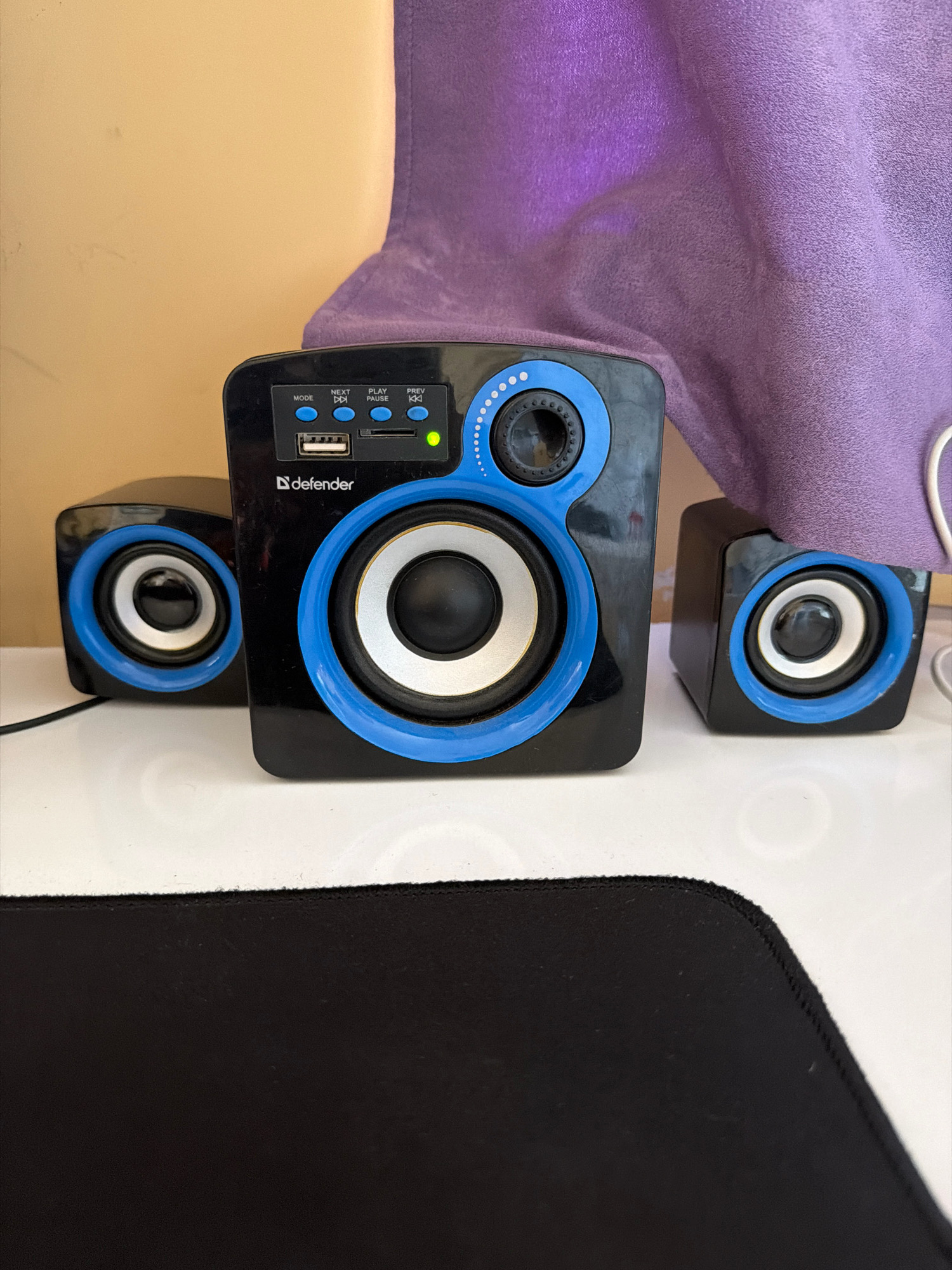 A black Defender brand speaker system with blue accents is displayed on a white surface. The central speaker, which is larger, features a USB port, SD card slot, and several control buttons labeled "MODE", "NEXT", "PLAY/PAUSE", and "PREV". A green indicator light is illuminated. To the left and right of the central speaker are two smaller, matching satellite speakers. A purple cloth is draped behind the speakers, and a tan wall is visible in the background. A black mousepad is in the foreground. There are no people or animals present in the image, and no activity is depicted. The location is not specified in the image or context.