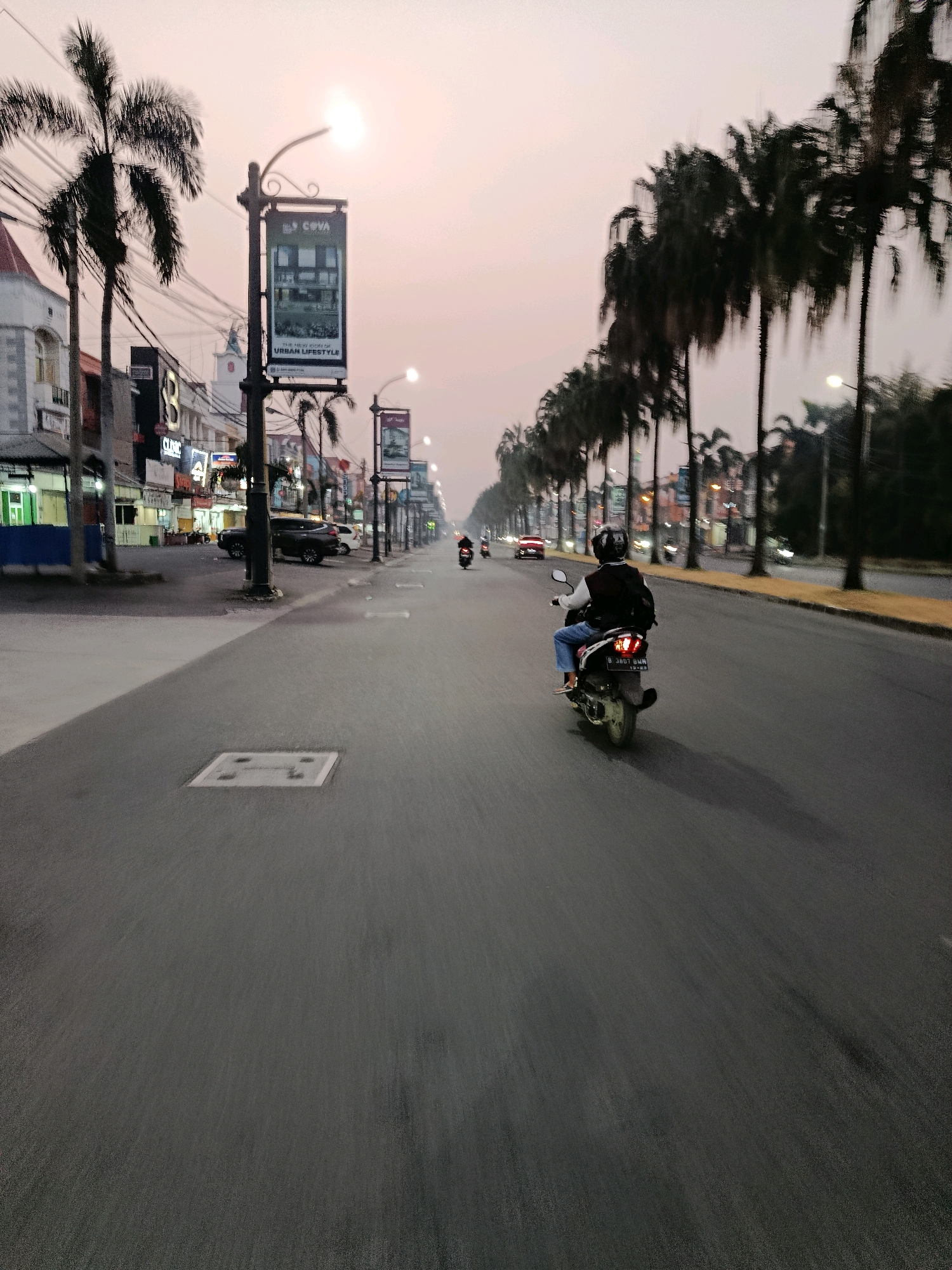 The image depicts a street scene in Bekasi, Indonesia, at dusk. A person on a motorcycle, wearing a helmet and a backpack, is riding away from the camera down a wide, paved road. The road is lined with palm trees and streetlights on both sides. On the left side of the road, there are buildings with storefronts, some of which are illuminated. Further down the road, other vehicles and motorcycles can be seen. The sky is a soft, hazy pinkish-orange, indicating the setting sun. The overall impression is that of a typical urban street during twilight hours.